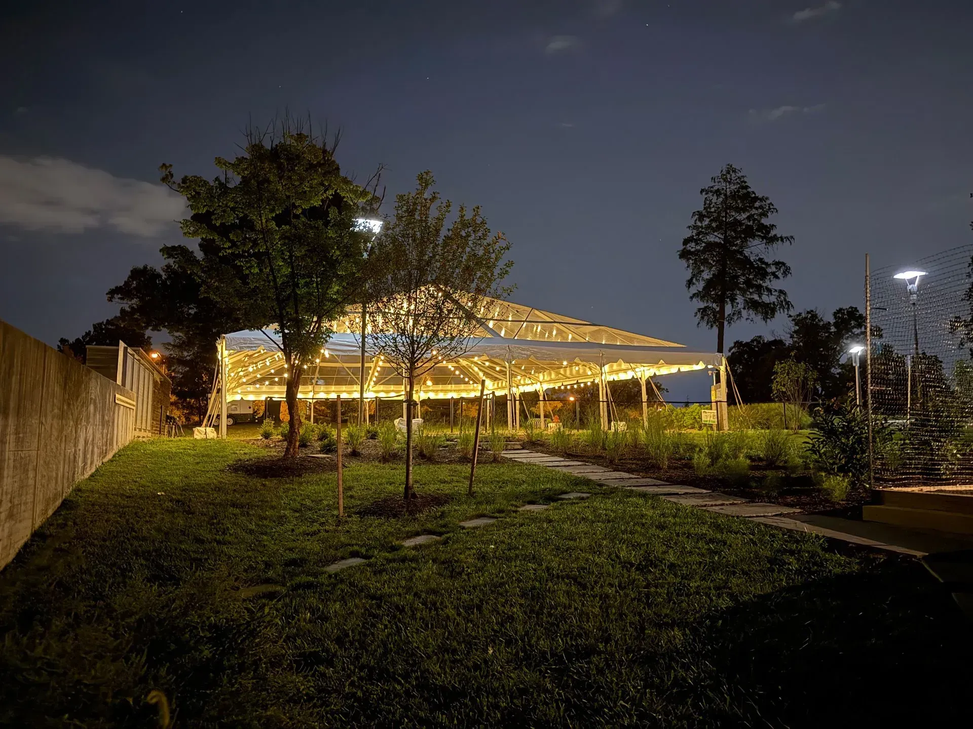 A lit canopy tent glows at night in a park with grass, trees, and stone pathways under a dark sky.