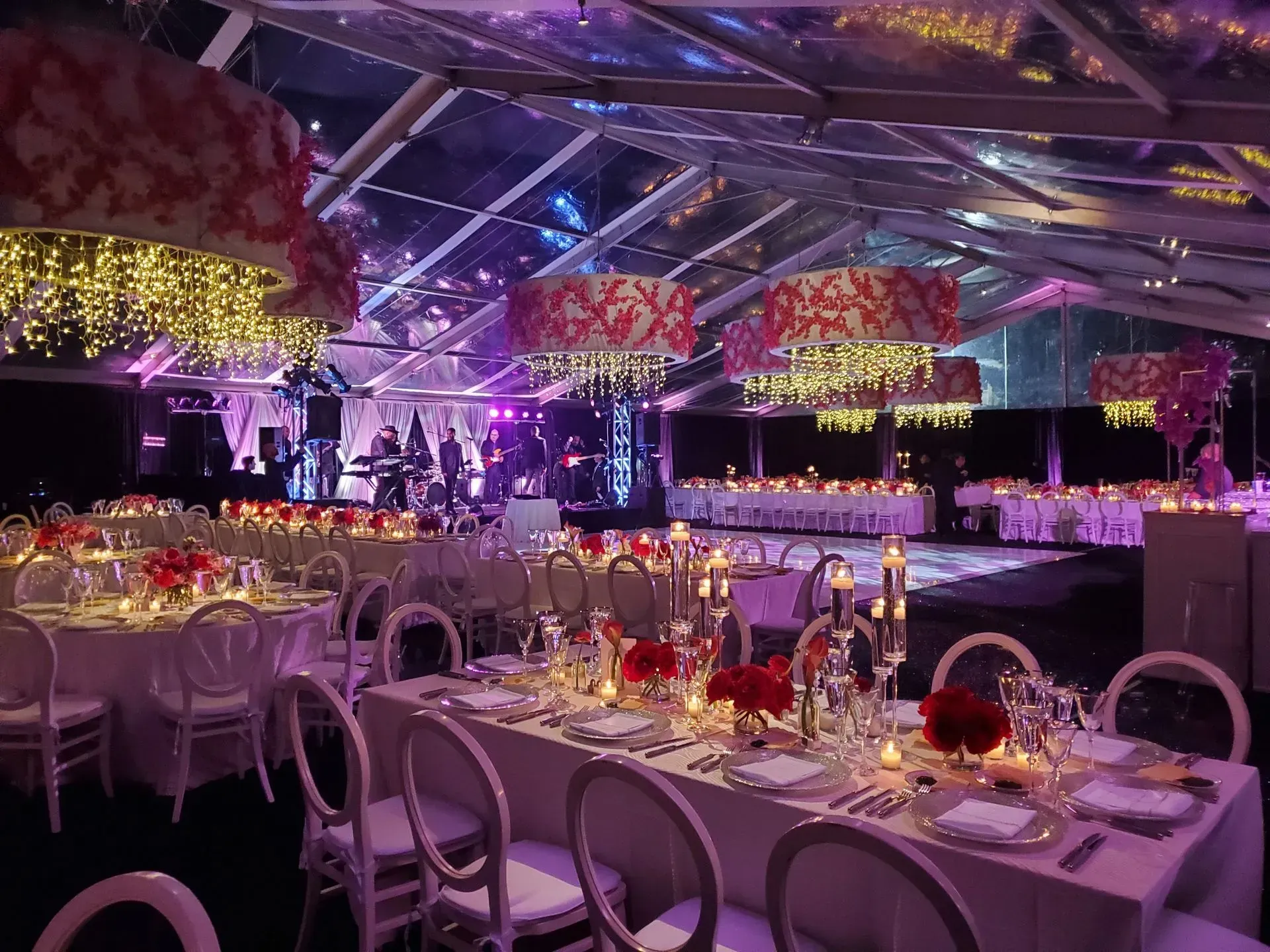 A formal event setup inside a clear-roofed tent with round and rectangular tables, red floral centerpieces, and lighting.