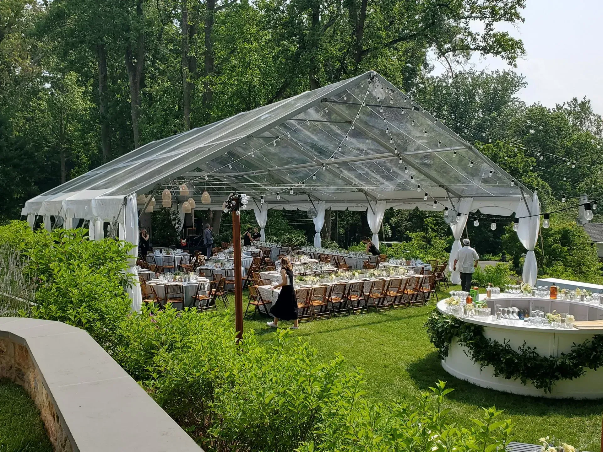 A clear-roofed event tent set up on a grassy lawn with tables and chairs, adjacent to a round bar decorated with greenery.