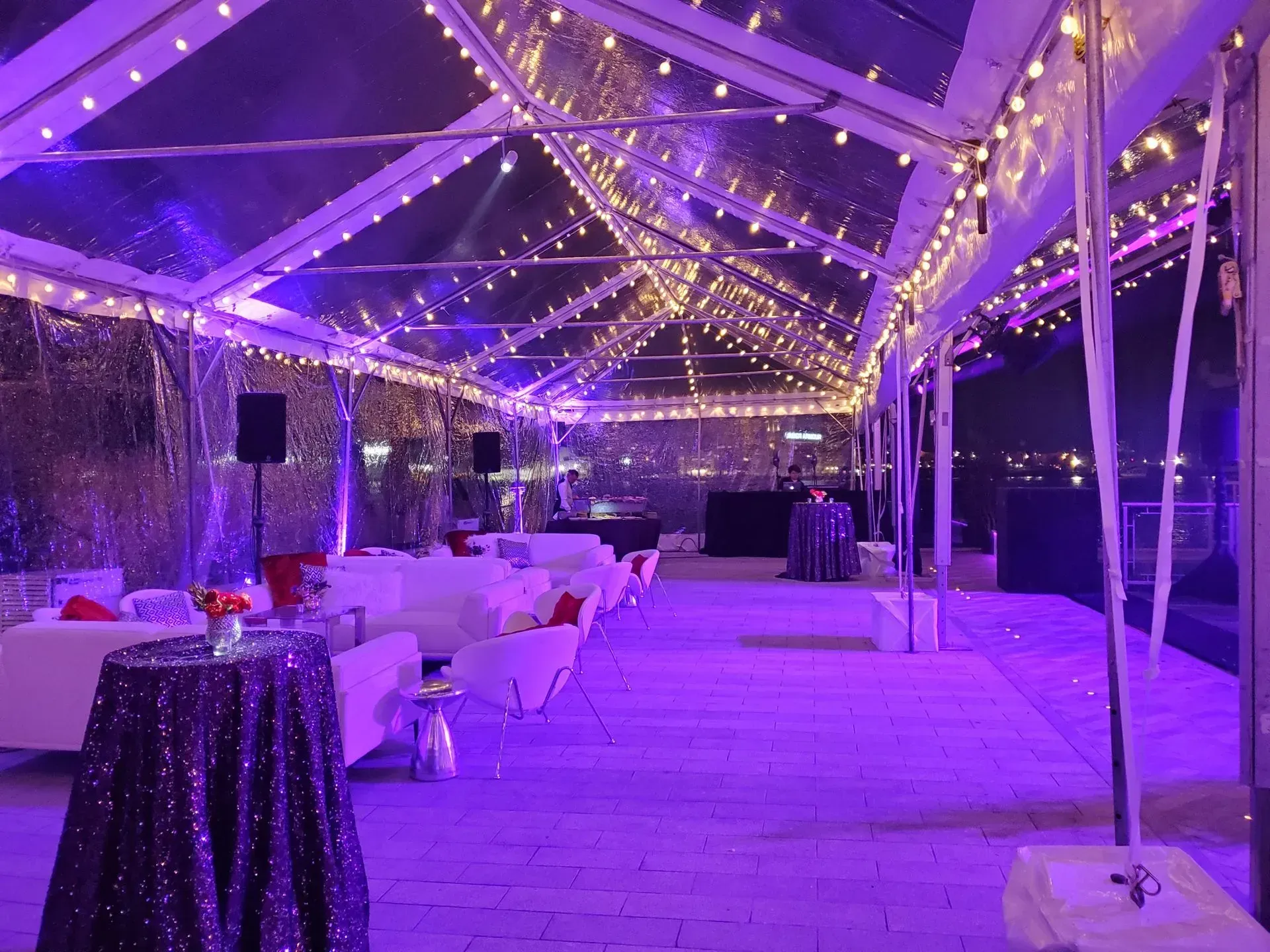 An outdoor event space under a clear-roofed tent at night, decorated with purple uplighting and string lights.