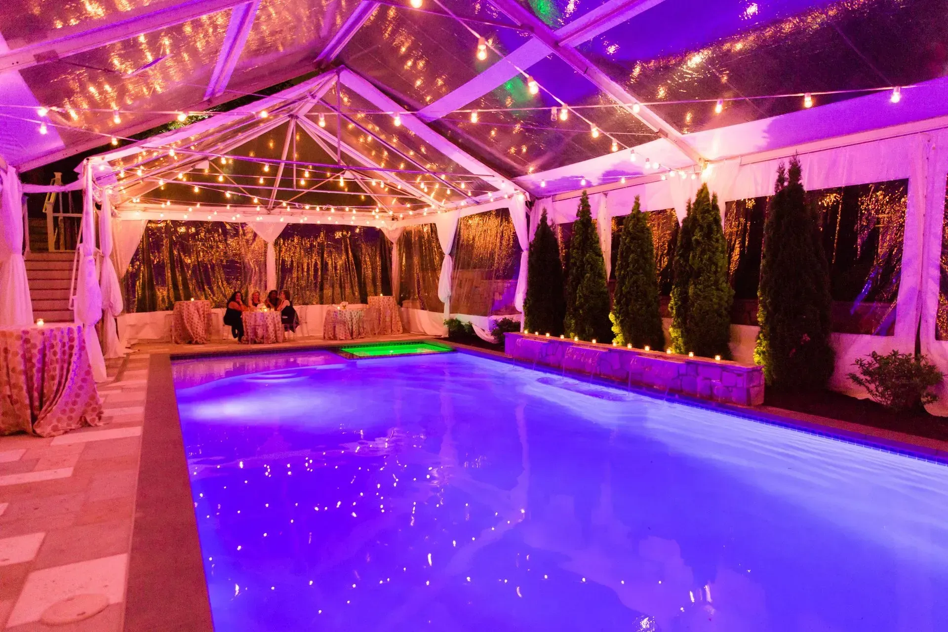 A pool glows with purple light inside a clear, string-lit tent, with evergreen trees and tables visible in the background.