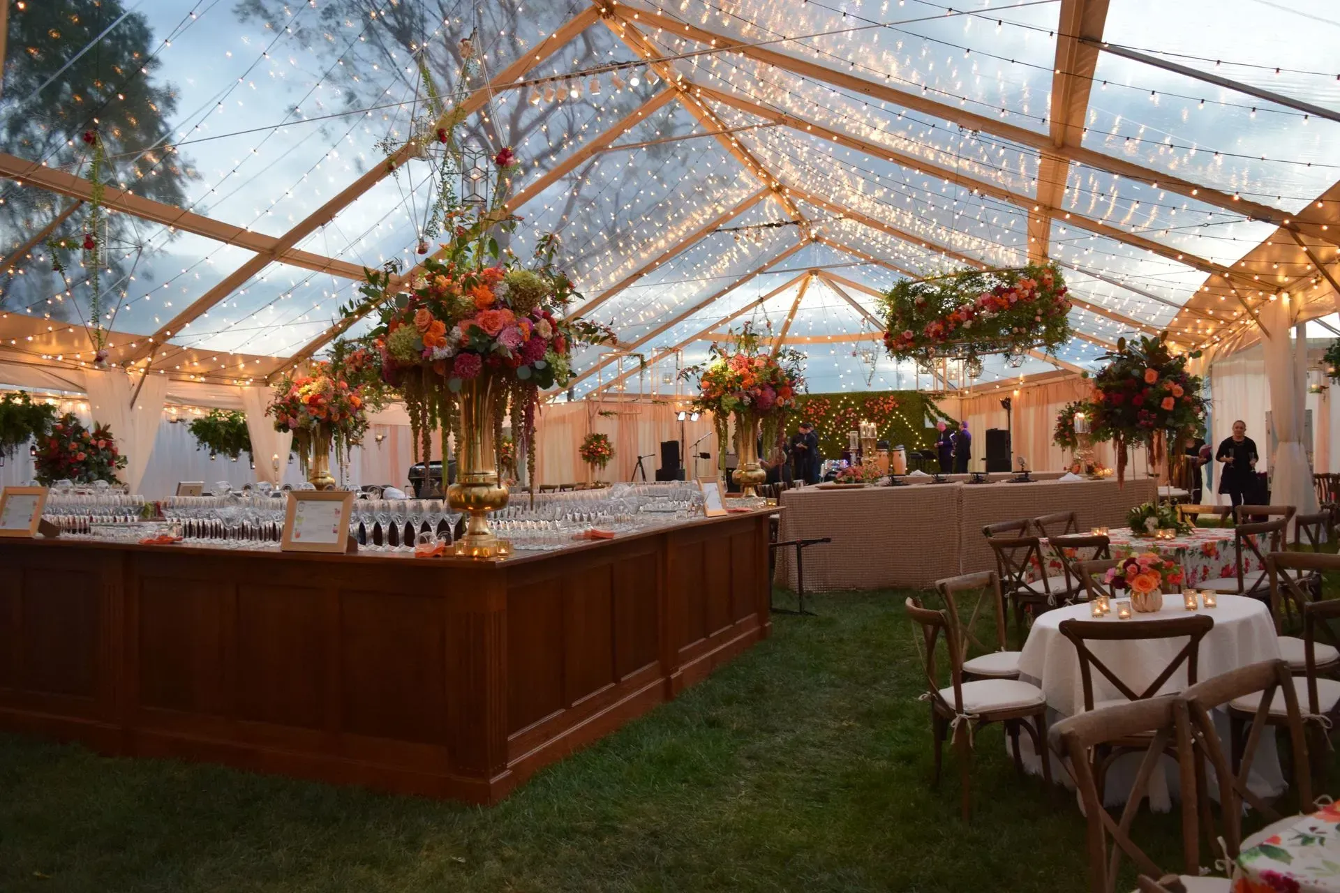 A festive outdoor event tent with a clear ceiling, string lights, floral centerpieces, a wooden bar, and guest tables.