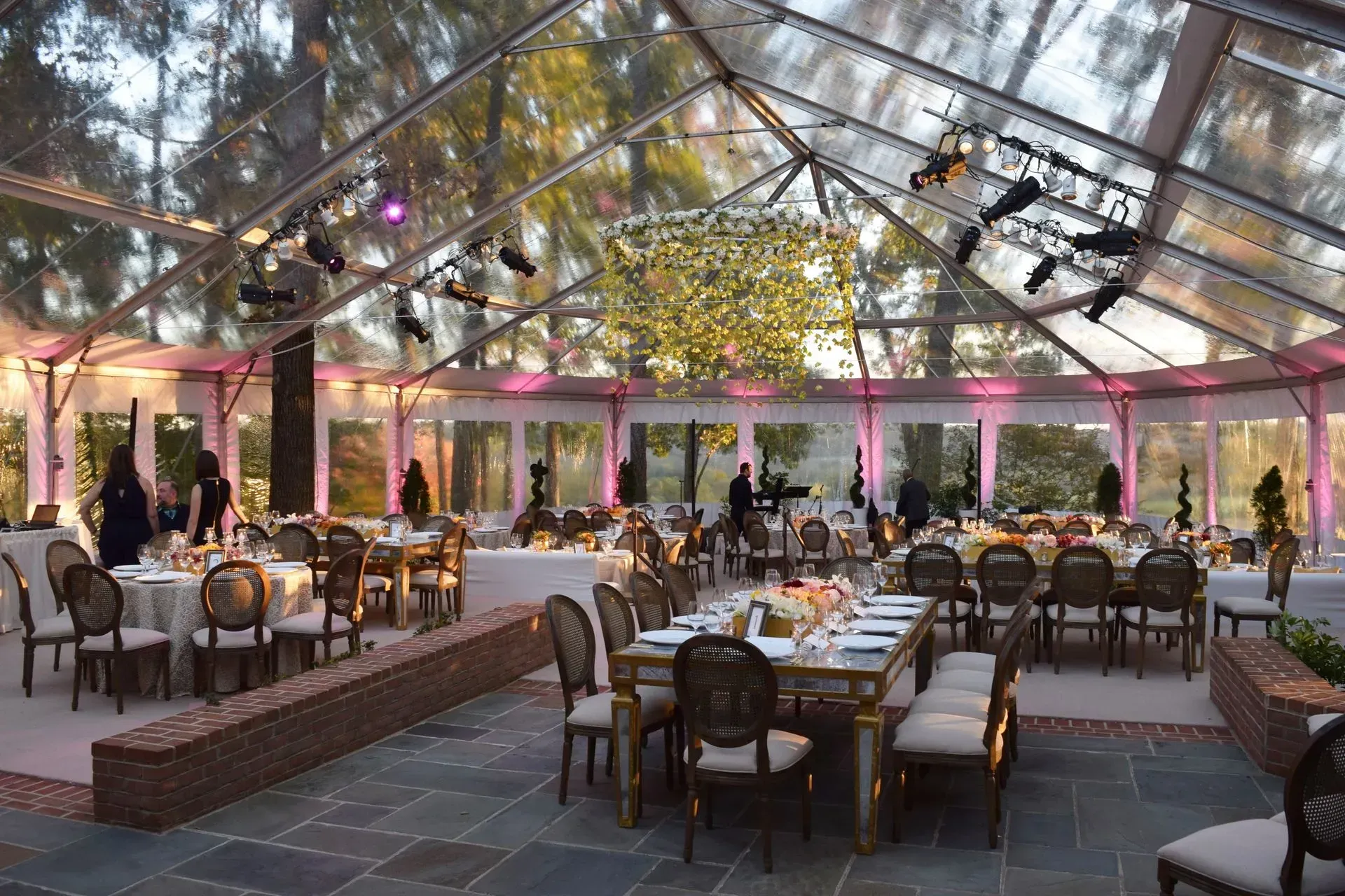 A clear-roofed event tent set up for an outdoor dinner party with elegant tables, chairs, and festive lighting.