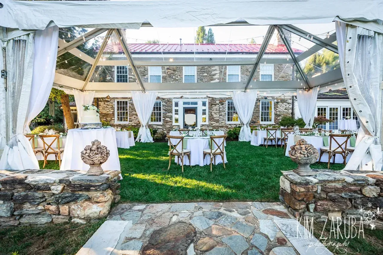 A clear event tent on a lush lawn, featuring white-clothed tables, chairs, and a stone house in the background.