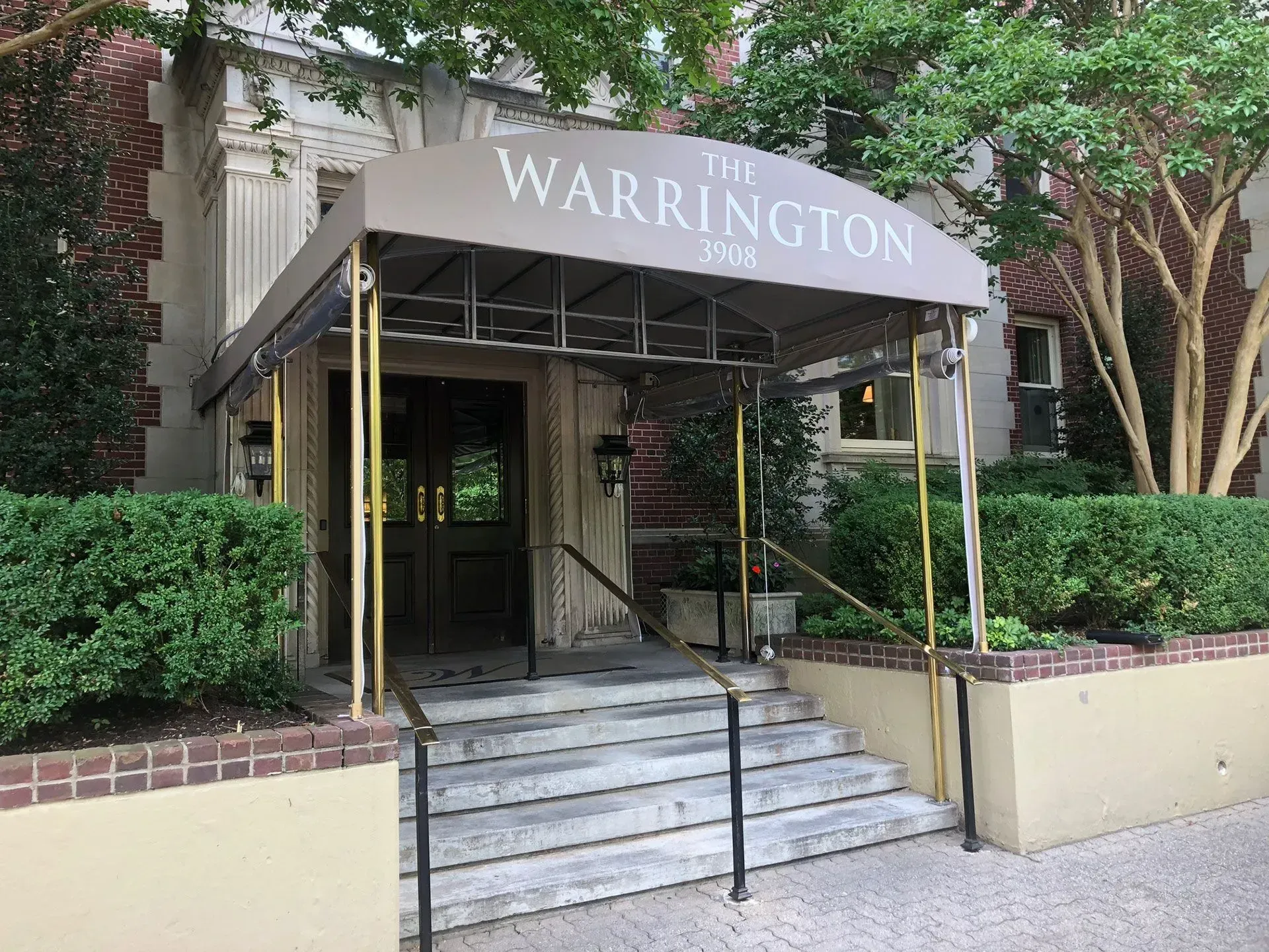 The Warrington apartment building entrance with a tan canopy, stairs leading to double doors, and green landscaping.