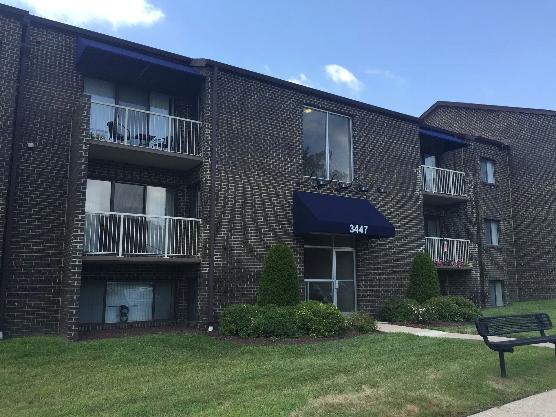 A three-story, brown brick apartment building with dark awnings, balconies, and a bench on a grassy lawn.