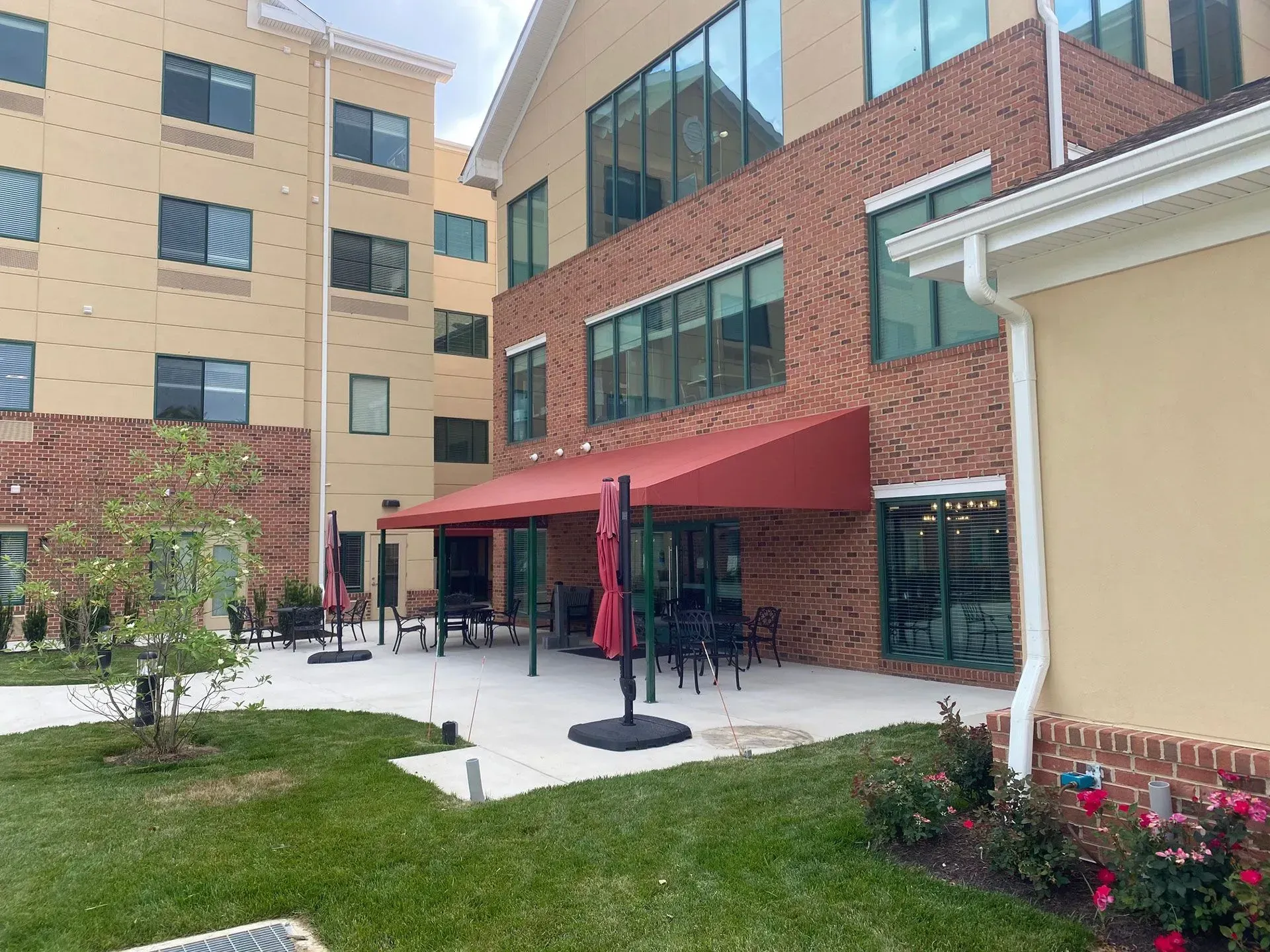 A red awning shades an outdoor patio with tables and chairs between a modern brick building and a tan multi-story complex.