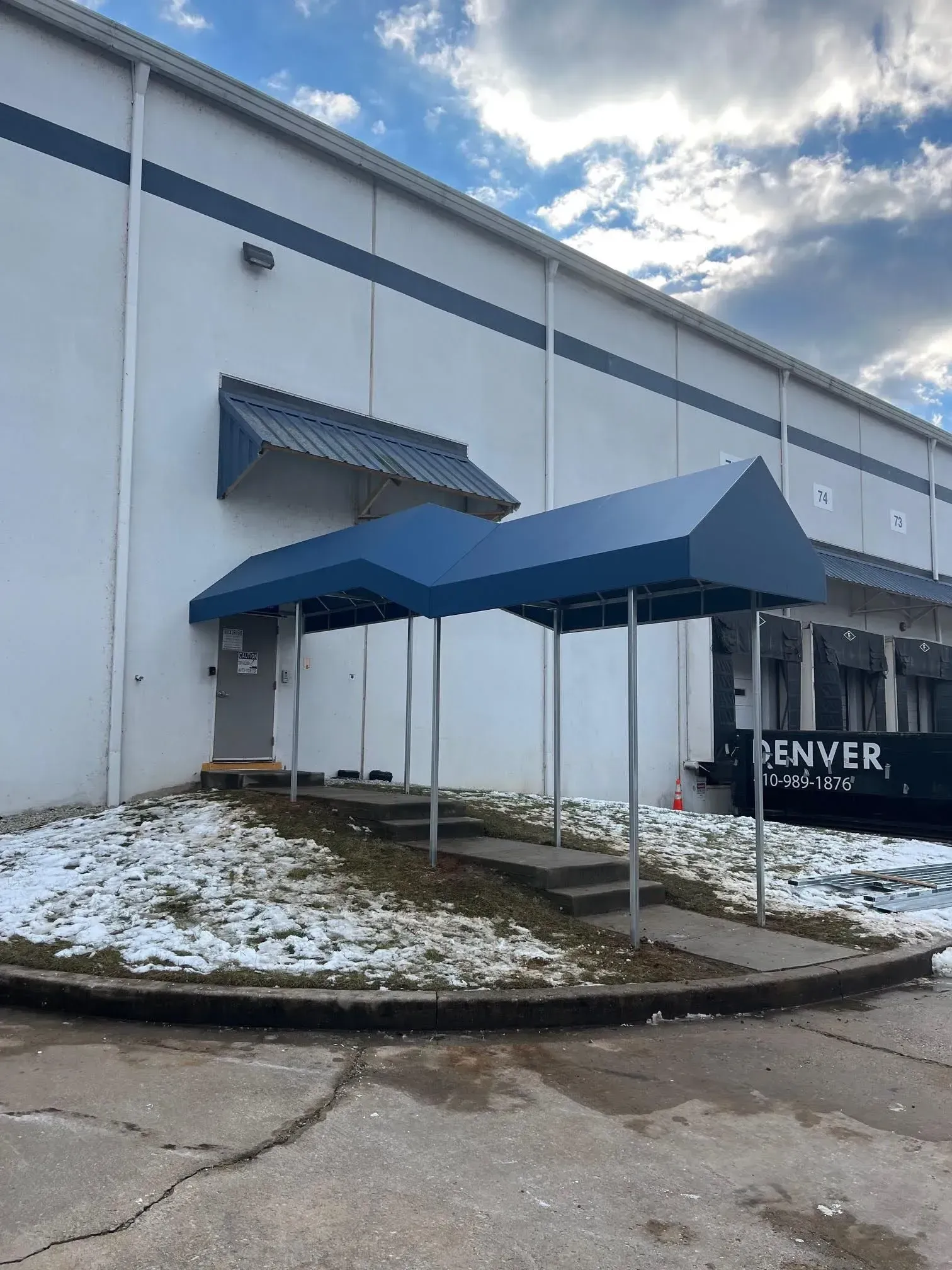 A white industrial building with a blue metal awning covering concrete steps leading to an entrance door.