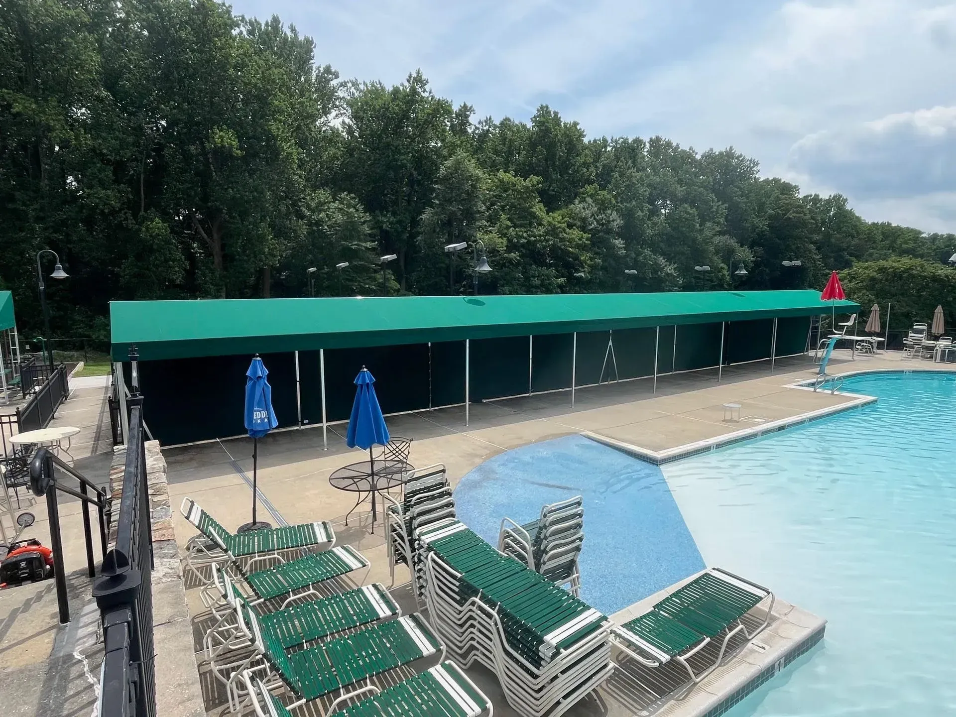A long green awning covers a patio next to a swimming pool with green lounge chairs and blue umbrellas on the deck.