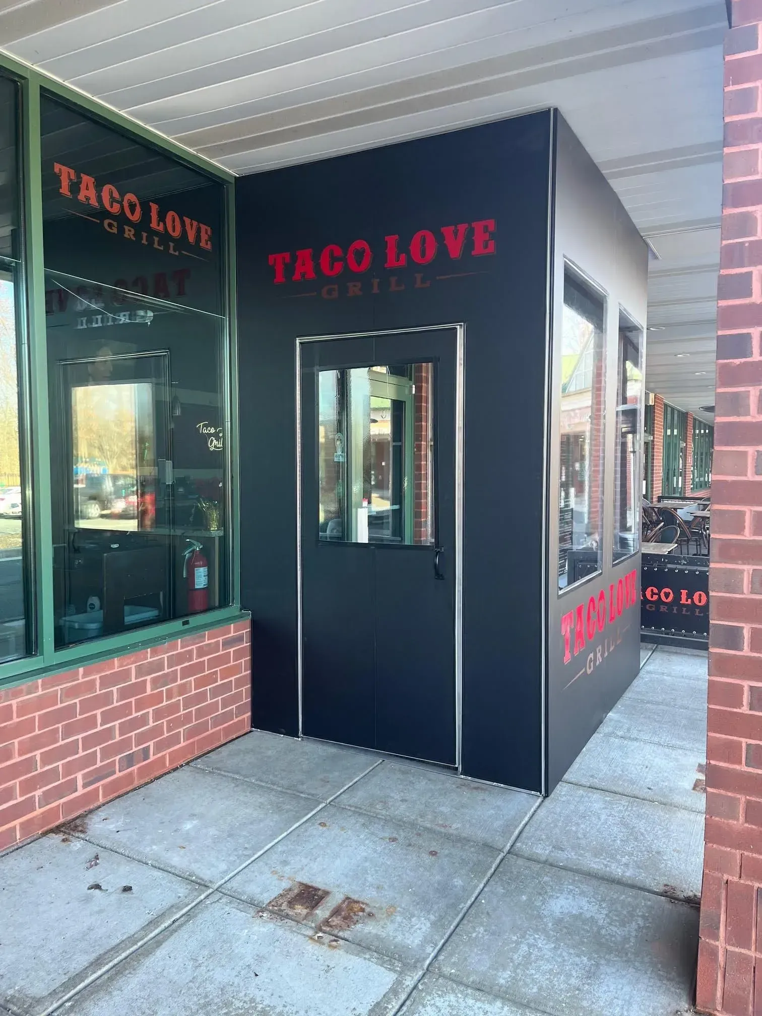 The exterior entrance of Taco Love Grill, featuring a black structure with red logo lettering and a glass door.