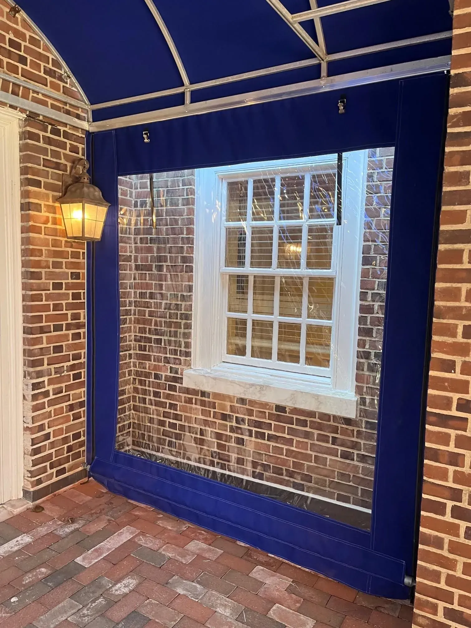 A brick building exterior featuring a window enclosed by a navy blue awning and clear protective curtain panels.