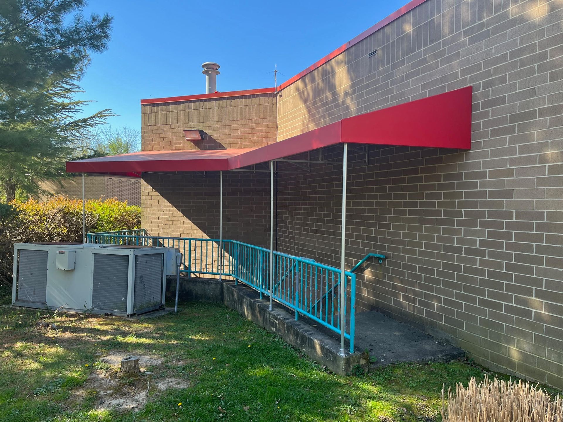 Brick building with red awning over a blue-railed ramp and outdoor HVAC units in a yard