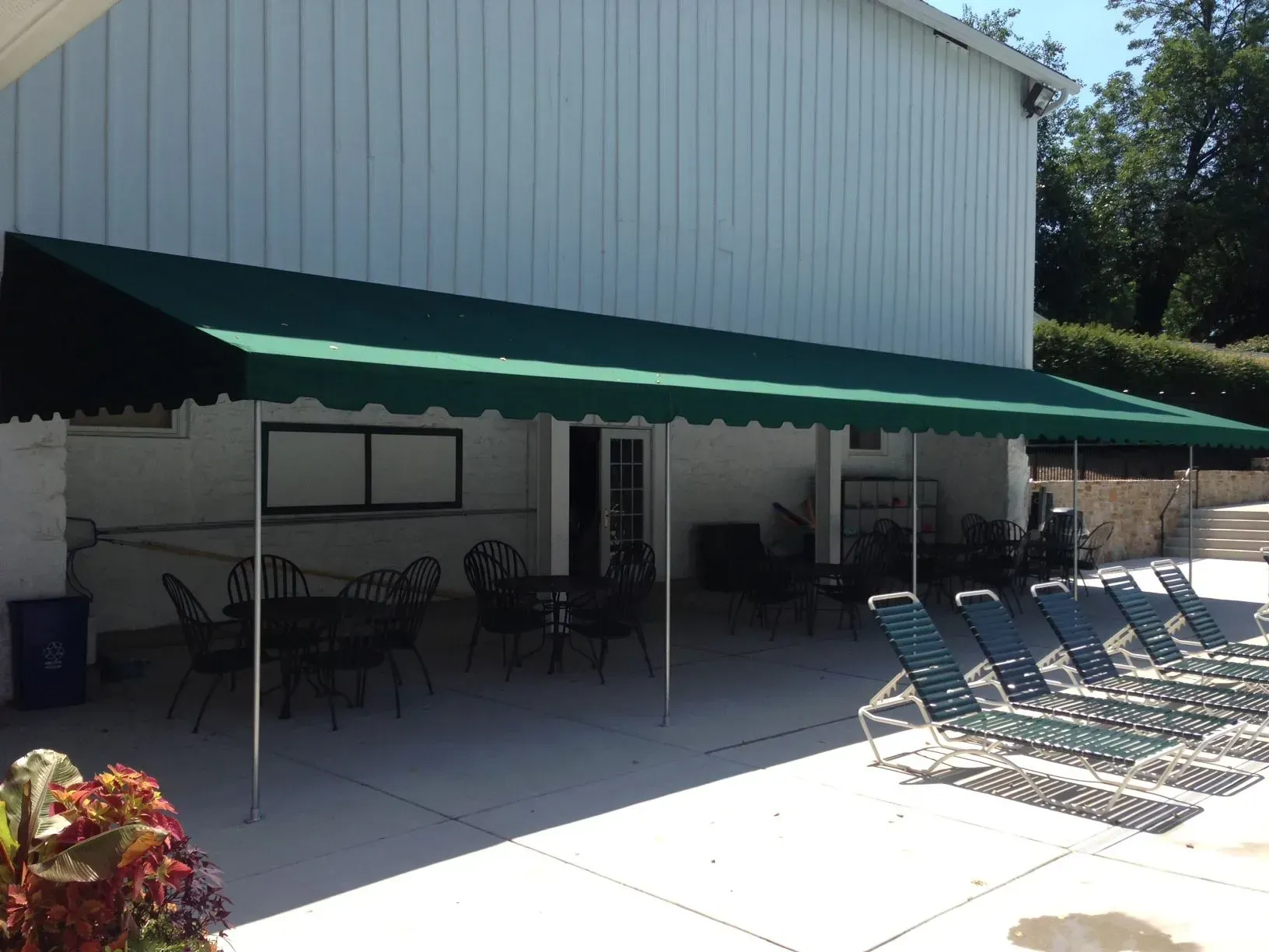 A long green awning covers a white building patio with outdoor tables and lounge chairs.