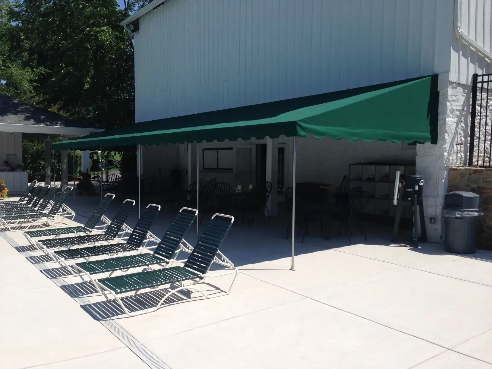 A row of dark green lounge chairs under a long, matching green awning on a sunny pool patio.