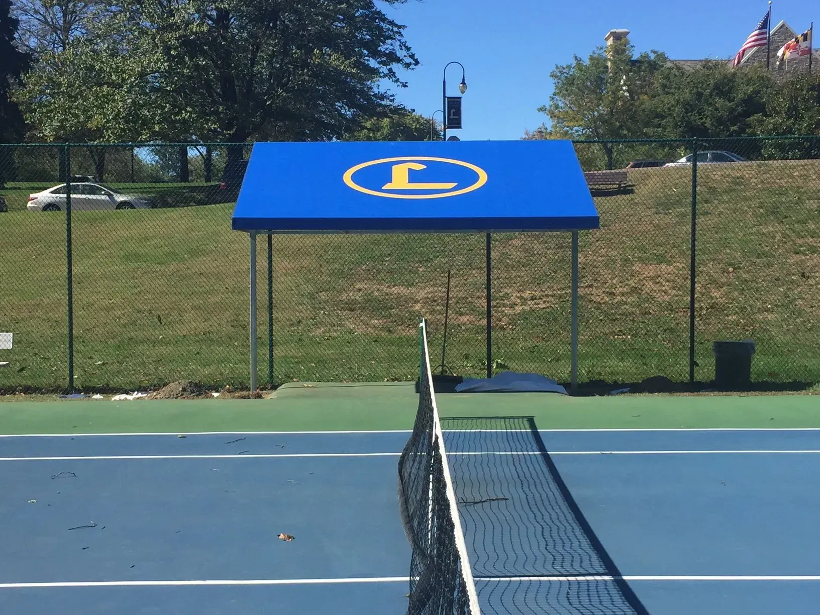 A blue canopy with a yellow letter L logo stands behind a tennis net on a blue and green outdoor tennis court.