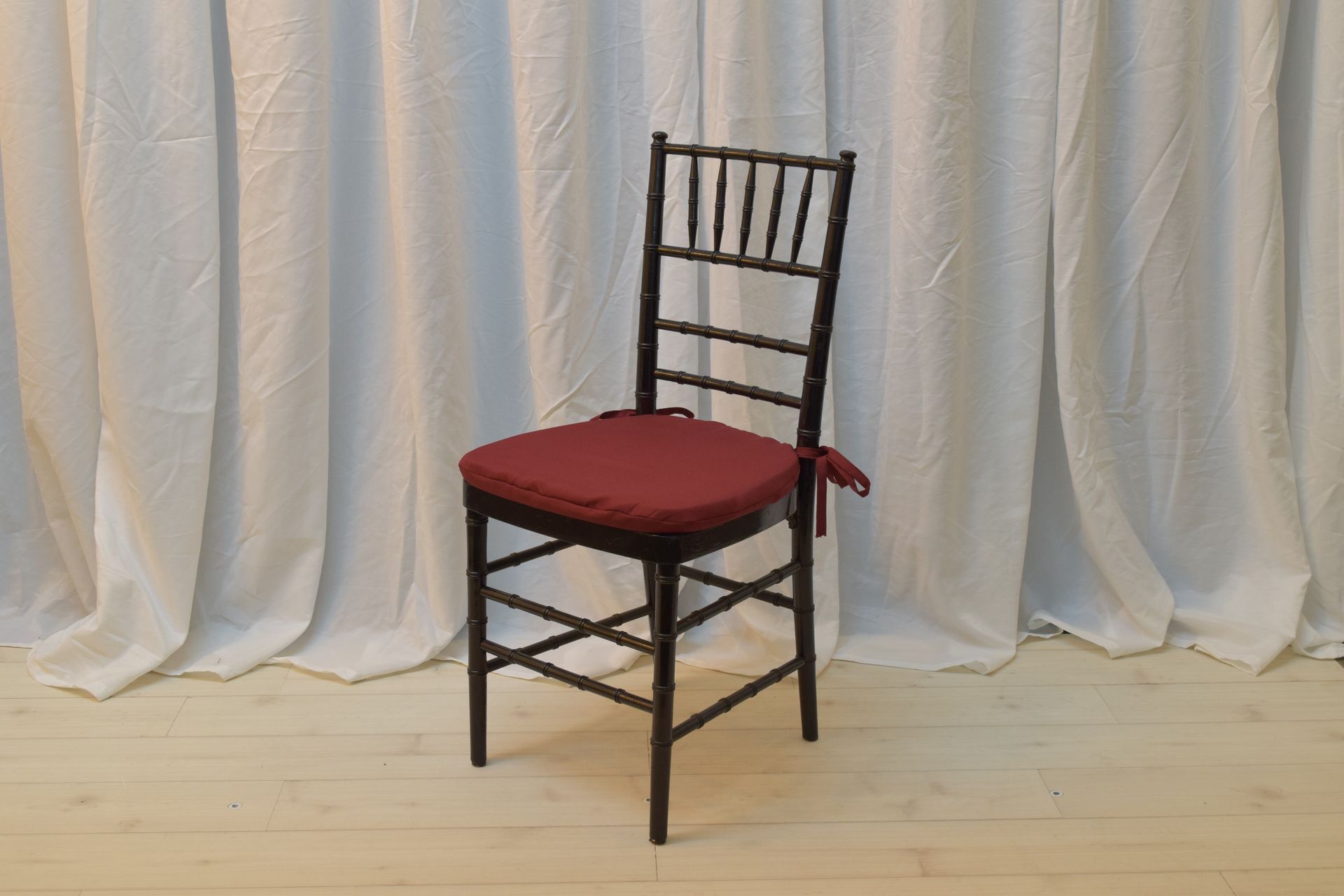 Black wooden chair with red cushion on a light wood floor in front of white curtains