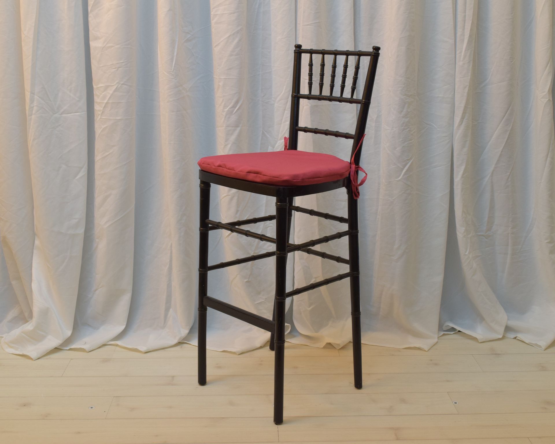 Black barstool with red seat cushion on a light wood floor in front of a white curtain