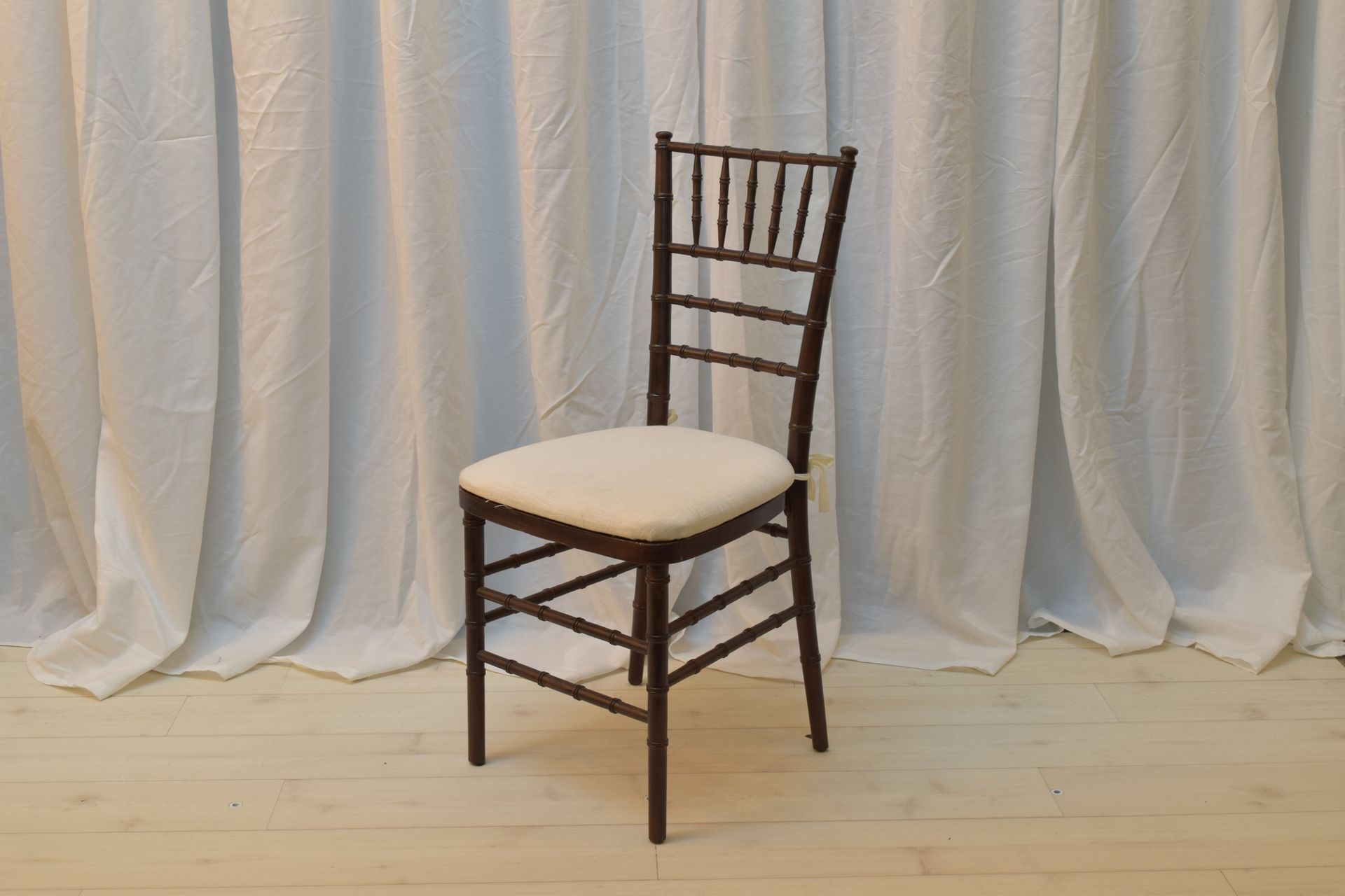 Brown wooden chair with beige cushion on a light wooden floor in front of white curtains