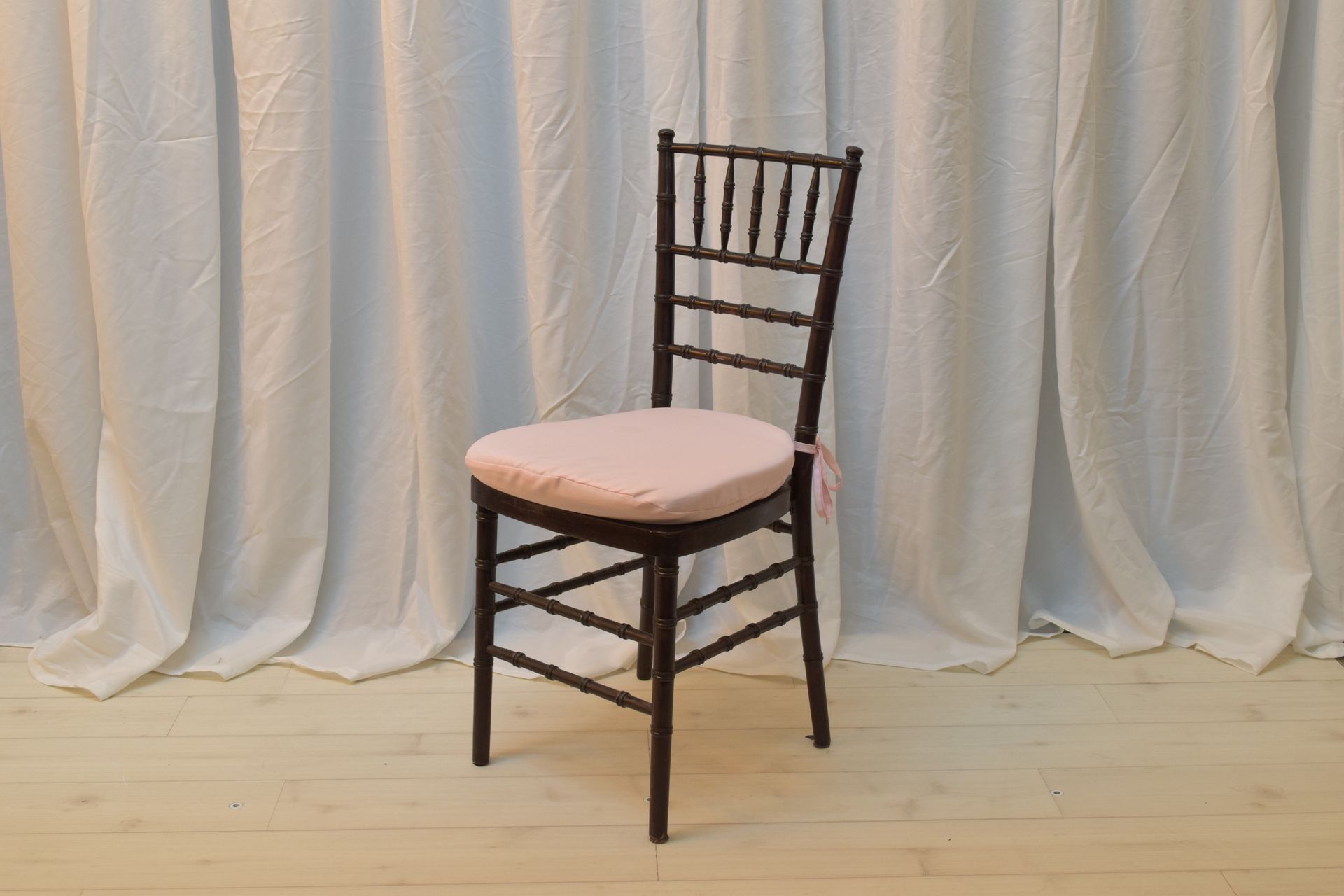Dark wooden chair with a pale pink cushion on a light wood floor in front of white drapes