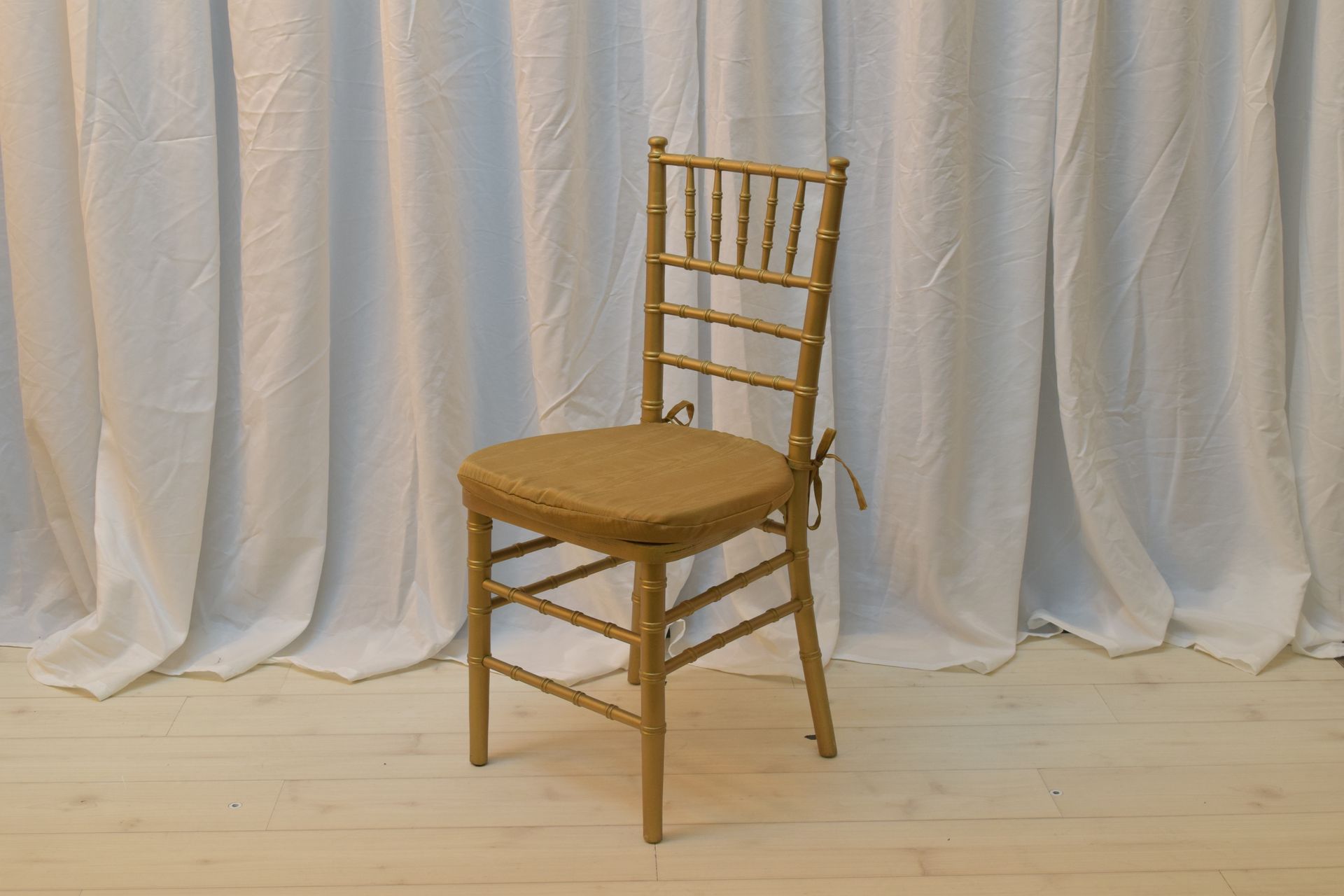 Gold chiavari chair with tan cushion on a light wood floor before white draped curtains