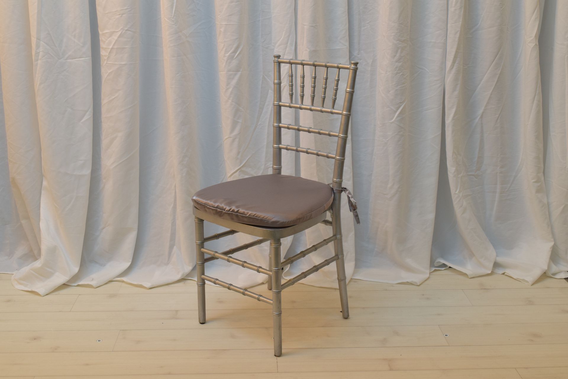 Gold chiavari chair with brown cushion on a light wooden floor against a white draped backdrop