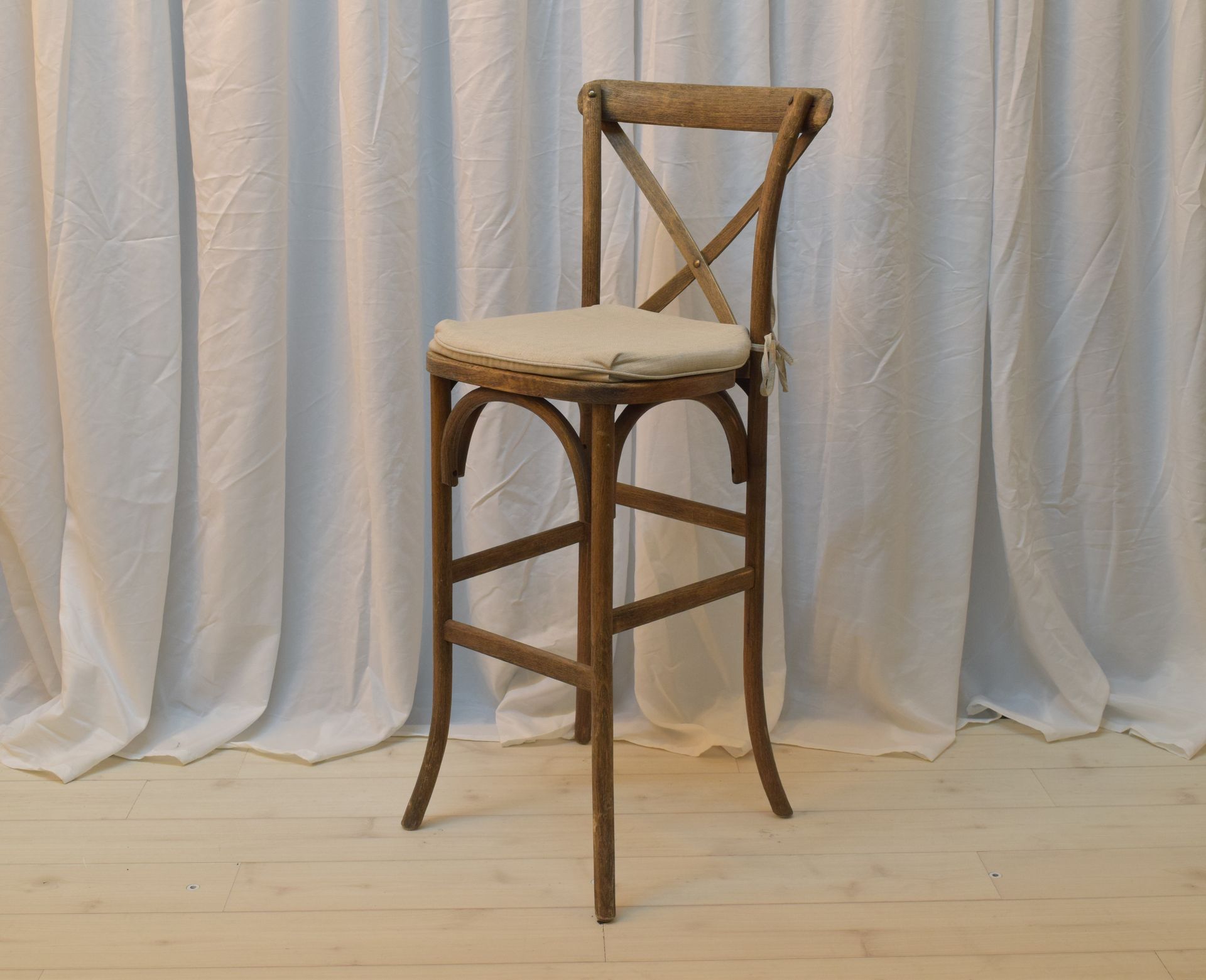 Rustic wooden cross-back bar stool with a light cushion on a pale wood floor, against white drapes