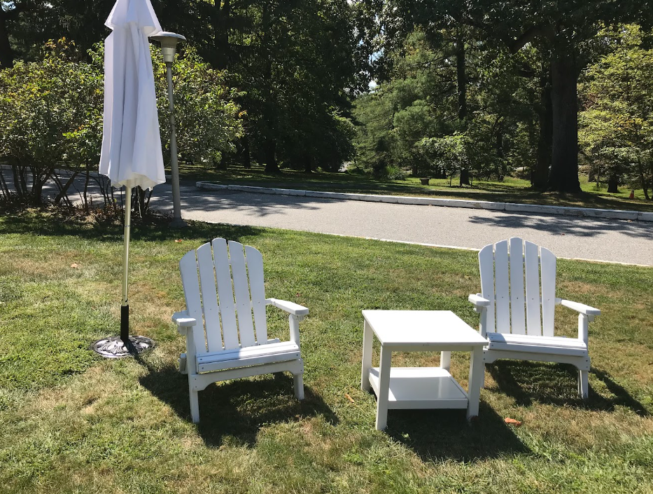 Two white Adirondack chairs and a small table on a sunny lawn beside a closed umbrella