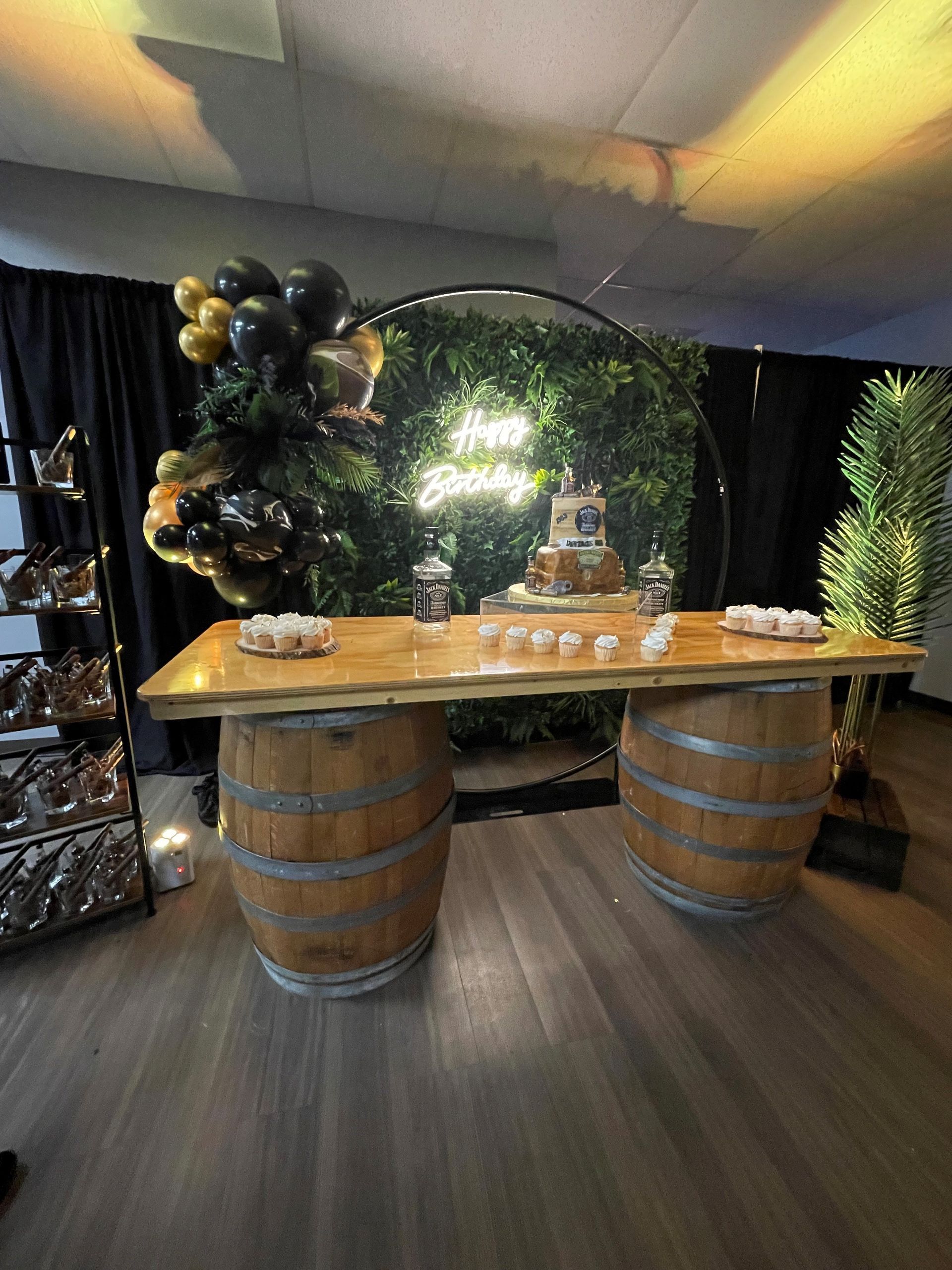 Rustic wooden table with barrel legs in a decorated event space, with black balloons and greenery backdrop.