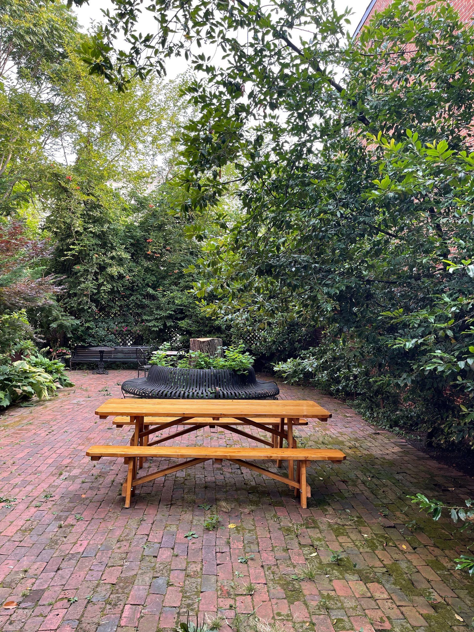 Wooden picnic table on a brick patio surrounded by lush green trees and shrubs.