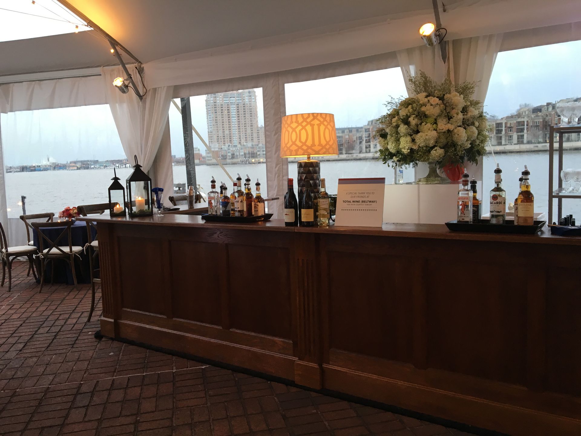 Open-air bar under a white tent by the waterfront, with bottles, lamps, and floral arrangements on a wooden counter