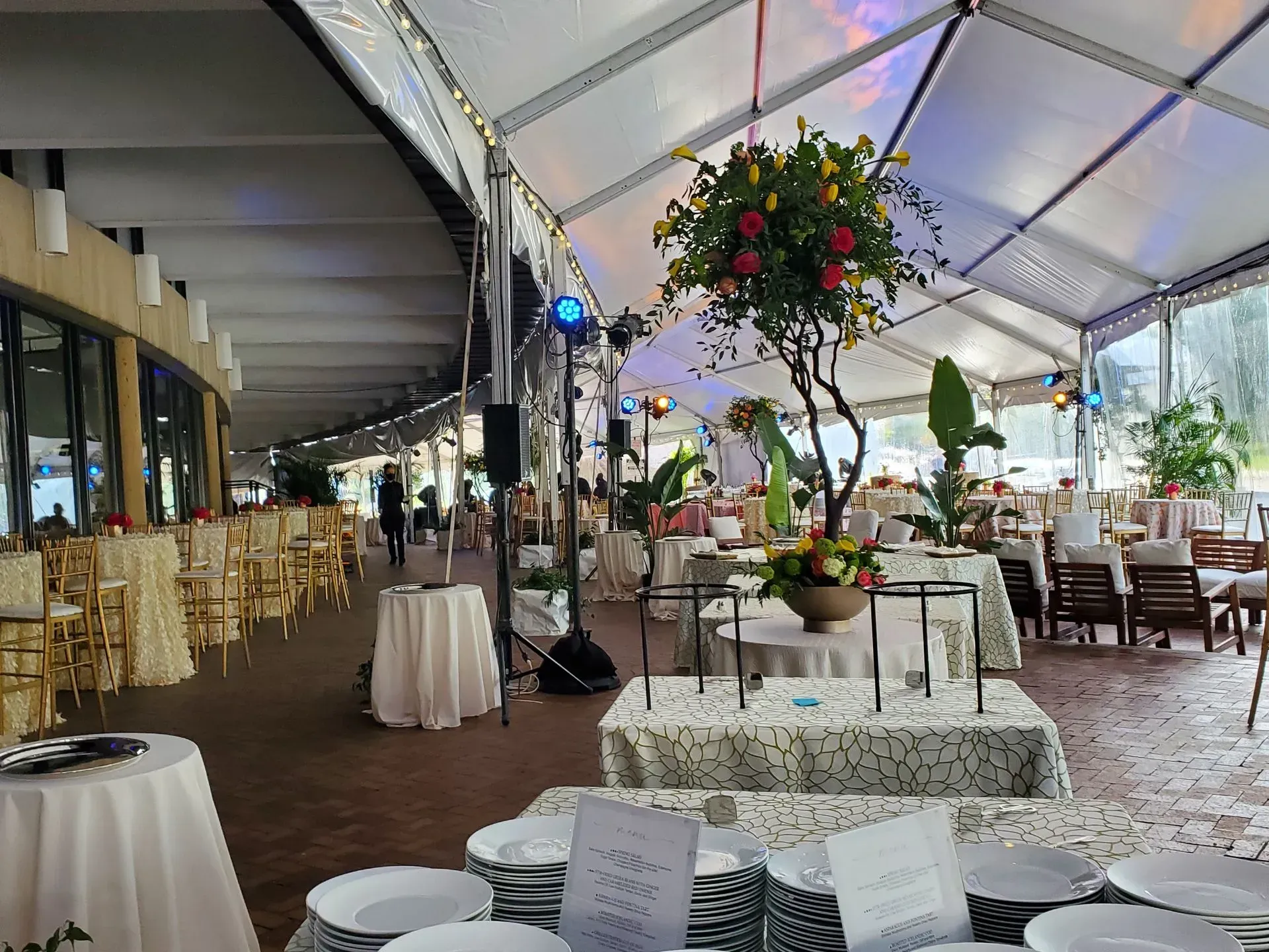 An event tent set for a catered gathering, featuring multiple tables with white linens, floral centerpieces, and plates.