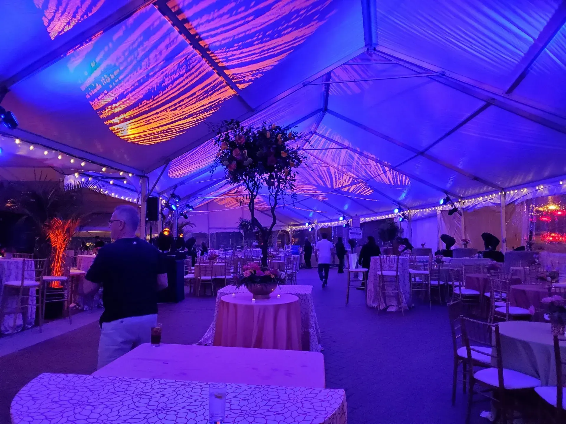 A tented event space illuminated in blue, featuring round tables with cream linens and string lights under the canopy.