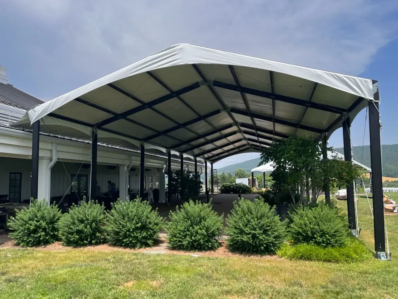 A large, rectangular white event tent with a black metal frame stands over a grassy lawn, near a building and shrubs.