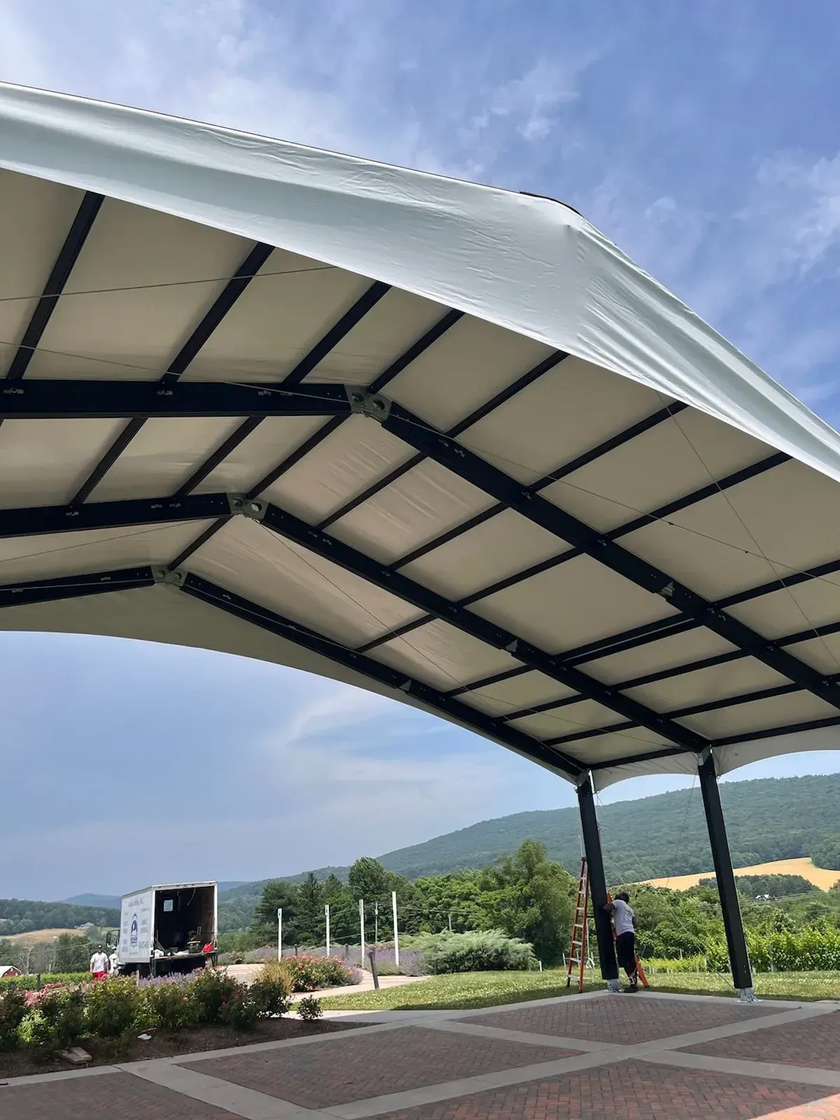 Workers set up a large, arched white event tent outdoors with mountains and a truck in the background.
