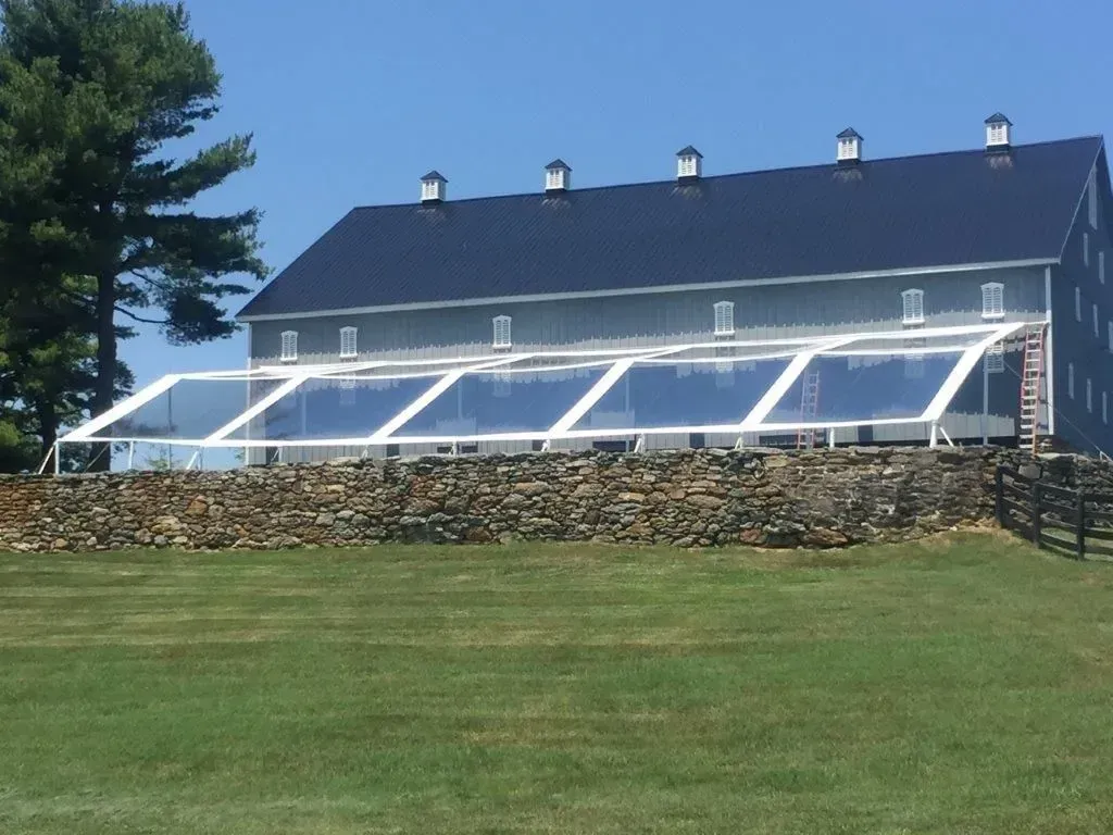 A large, grey barn with a dark roof sits behind a modern, clear-roofed event tent on a stone wall overlooking a green lawn.