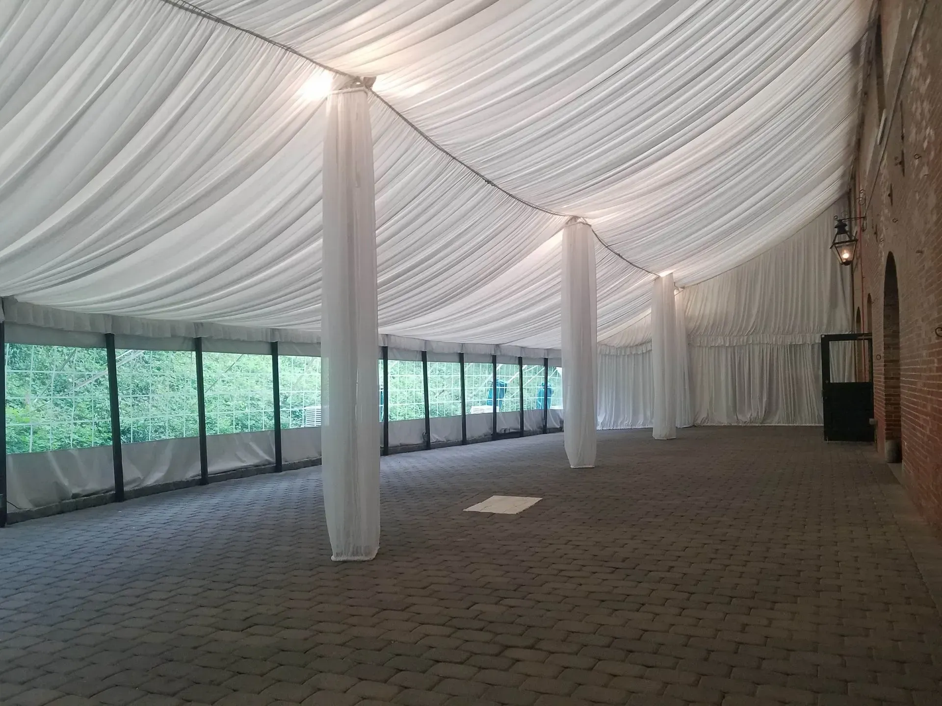 A long indoor event tent with white draped fabric ceilings, columns, and paved flooring, with views of trees outside.