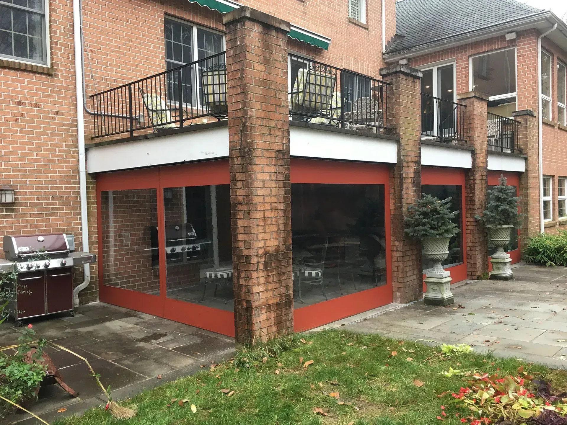 A brick house with a second-floor deck and an enclosed ground-floor patio featuring red panels and clear vinyl windows.