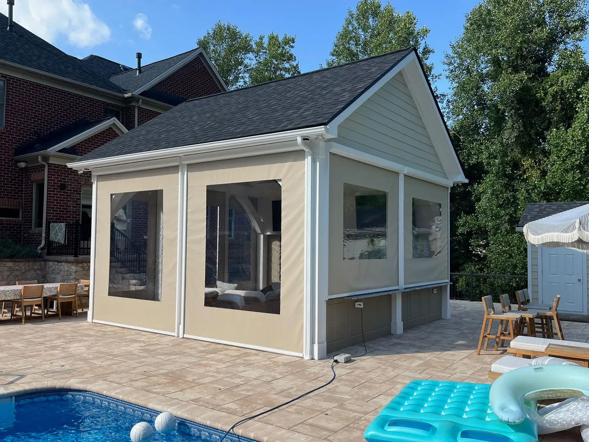 A poolside cabana with tan vinyl screens and clear windows, situated on a stone patio next to a house.