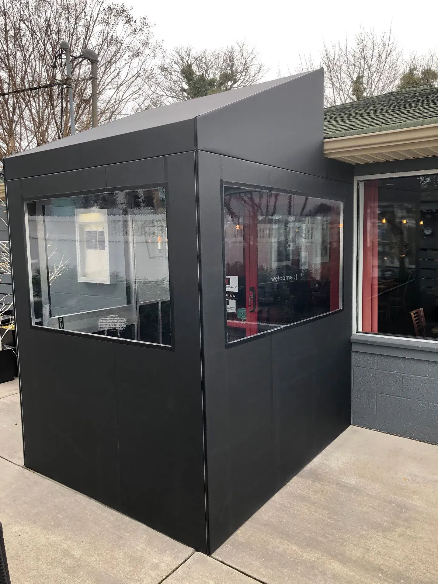 A black, angled outdoor enclosure with two large clear windows, attached to the side of a building on a concrete patio.