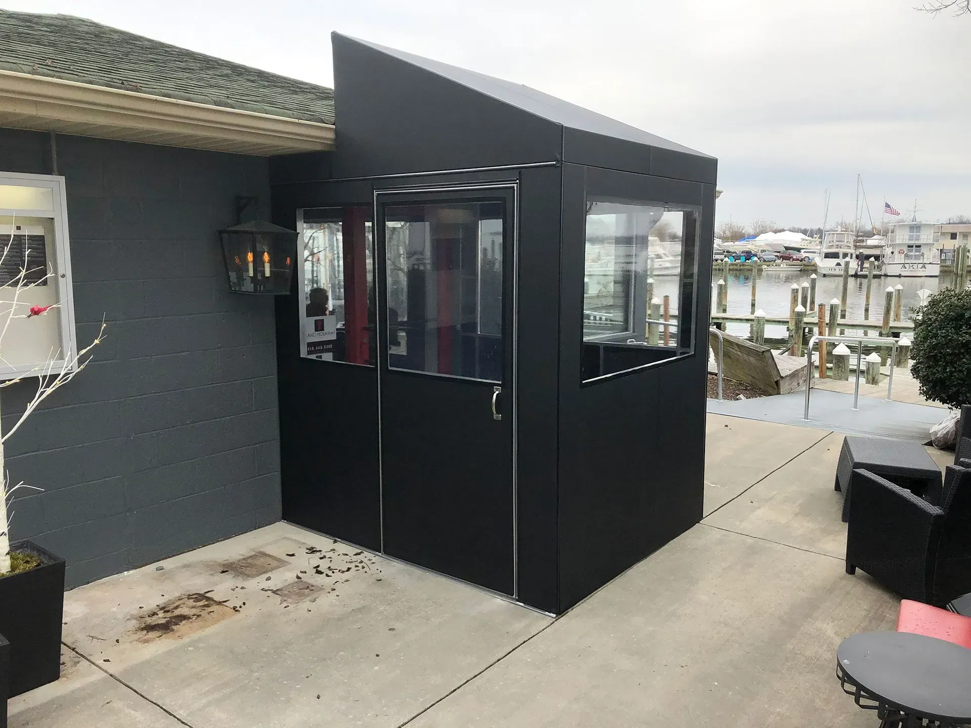 A black, enclosed outdoor patio vestibule with clear windows attached to the side of a grey building near a marina.