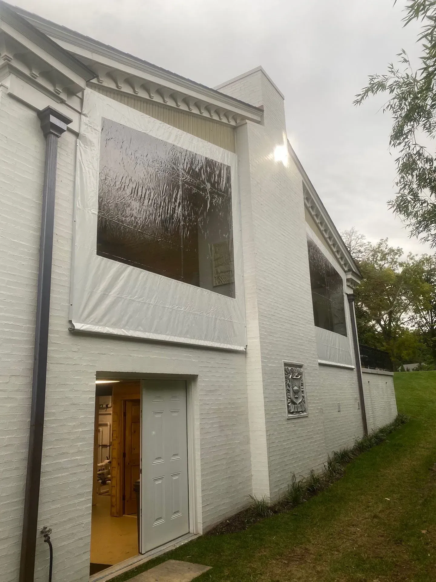 Exterior view of a white brick home showing clear vinyl patio enclosures on a raised deck and a basement doorway below.