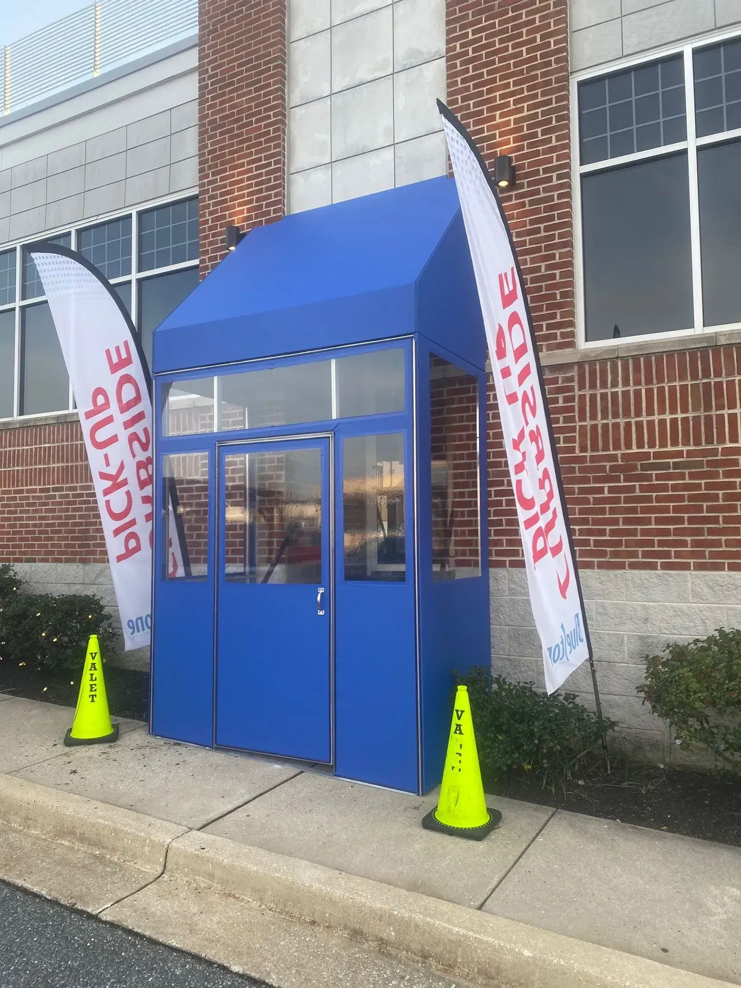 Blue curbside pick-up booth against a brick building, flanked by two branded vertical flags and bright yellow traffic cones.