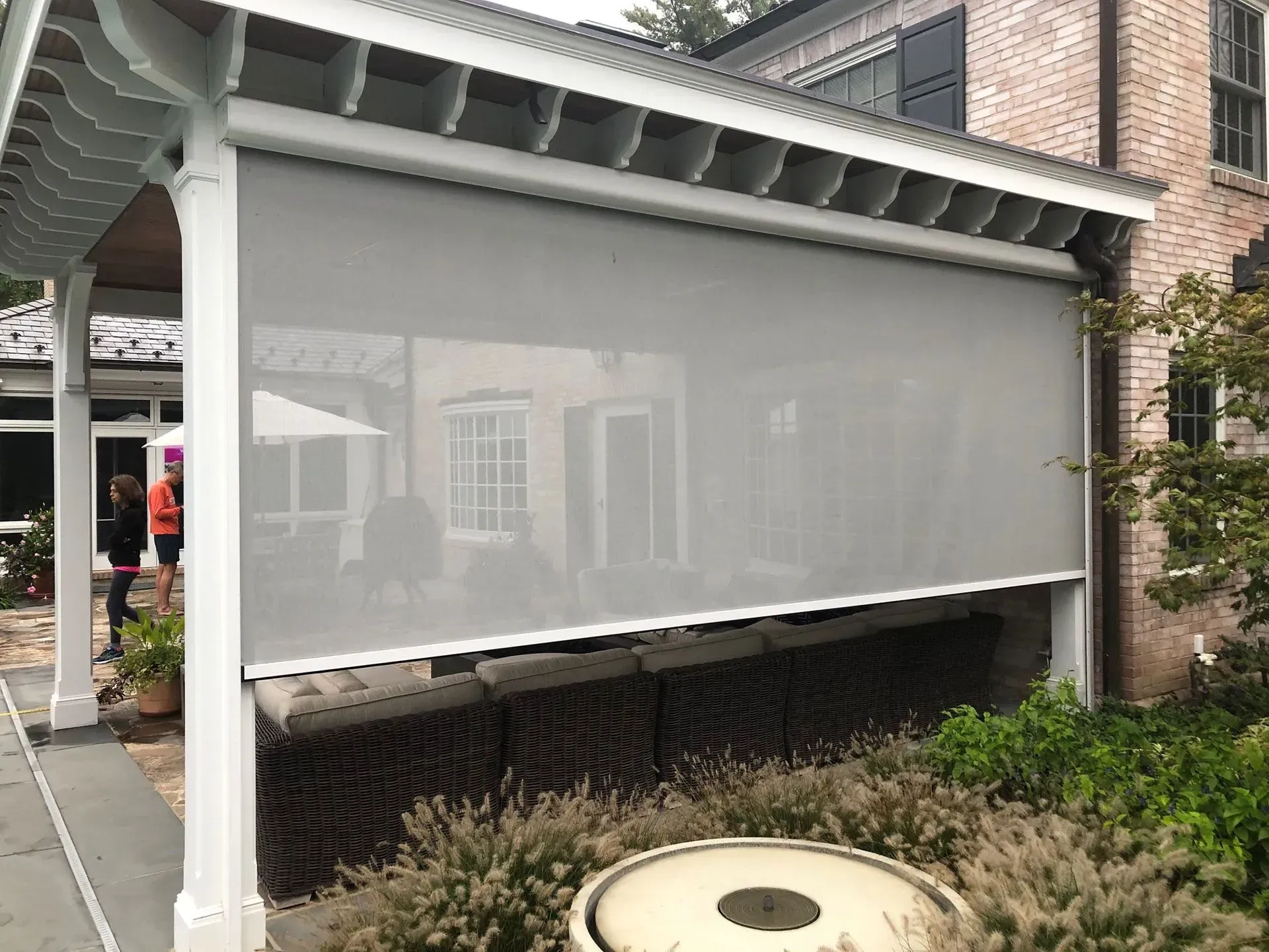 A light gray, semi-transparent solar shade hangs under a white wooden pergola attached to a brick house.