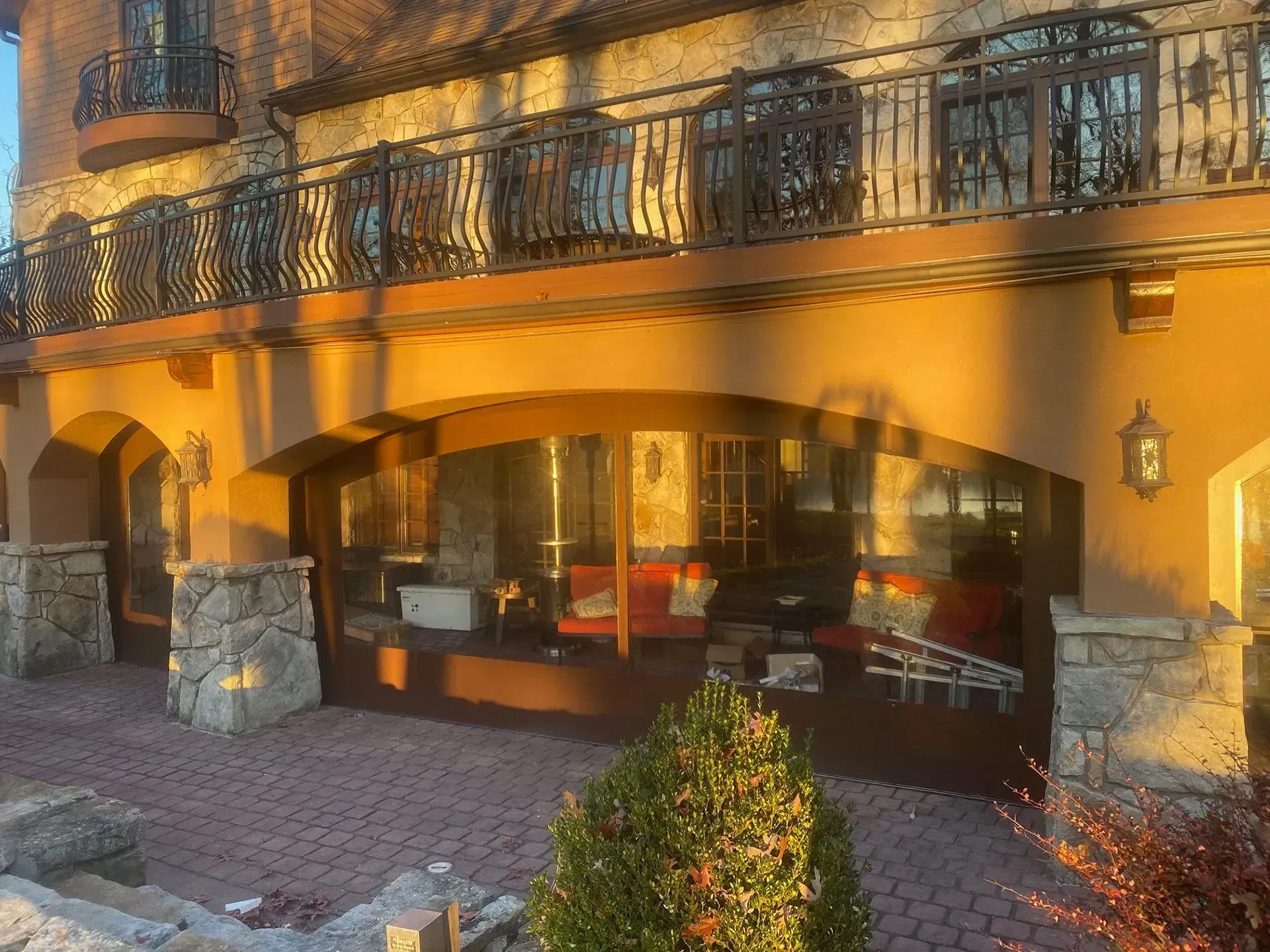 A sunlit stone building with an arched storefront window and a second-story metal balcony overlooking a paved patio.