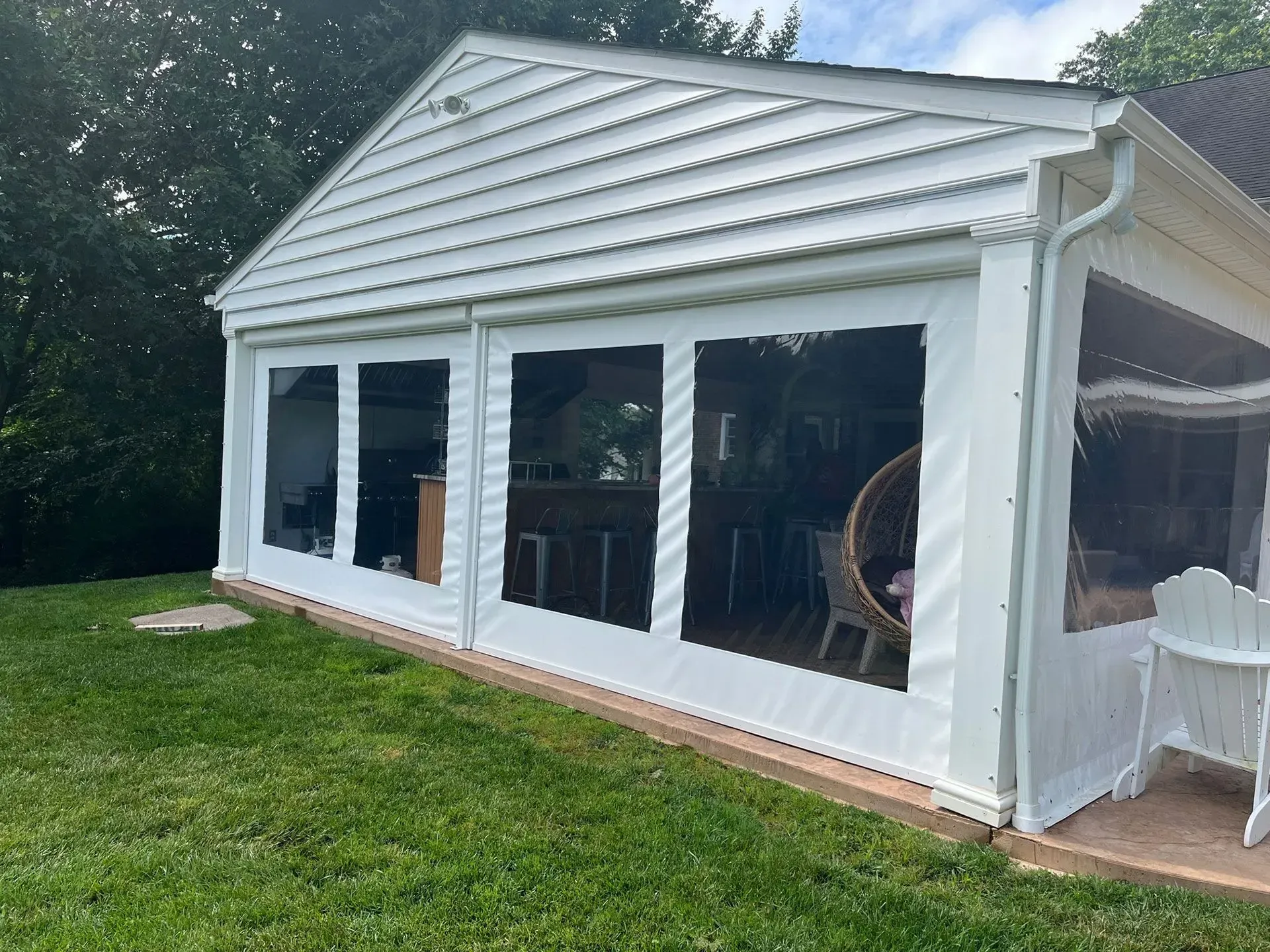 A white covered patio structure with clear plastic weather curtains, situated on a green lawn with outdoor seating inside.
