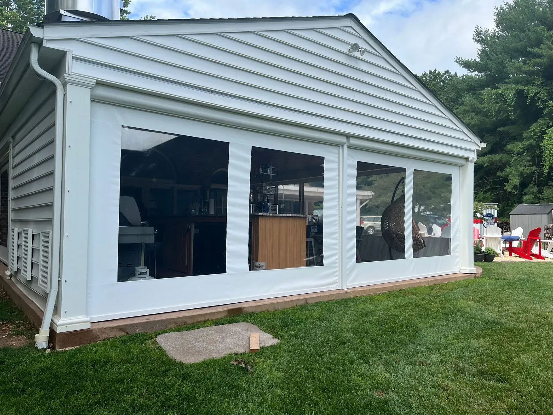A white outdoor structure featuring clear vinyl curtains, set against a green lawn with a red chair visible in the distance.