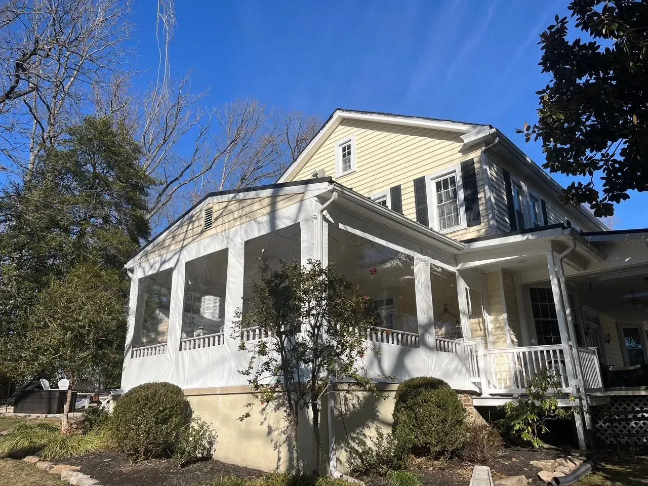 A two-story light yellow house with black shutters and a large, screened-in porch on the front, set under a blue sky.