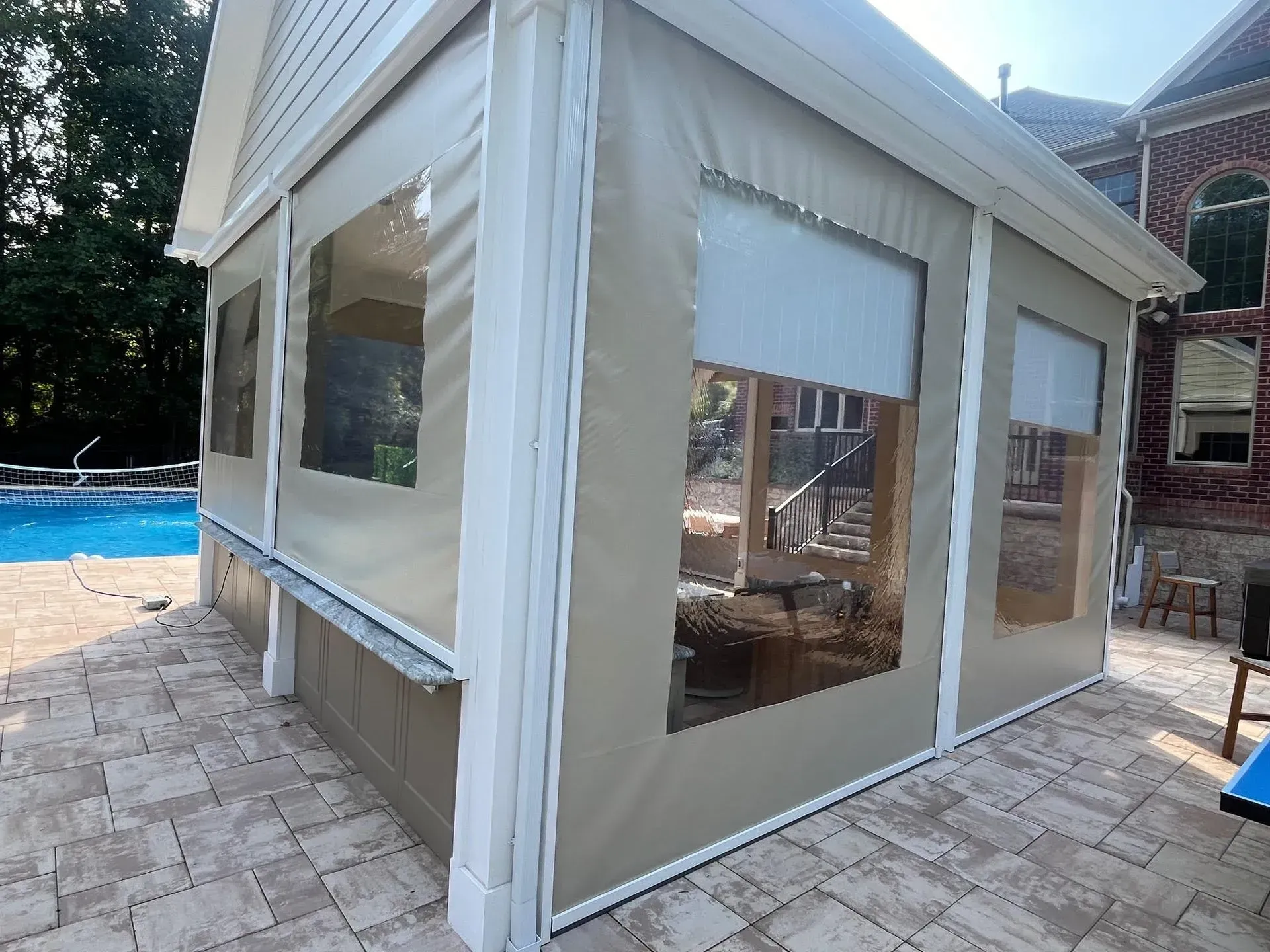 A beige outdoor enclosure with clear windows and white trim, installed on a stone patio beside a swimming pool.