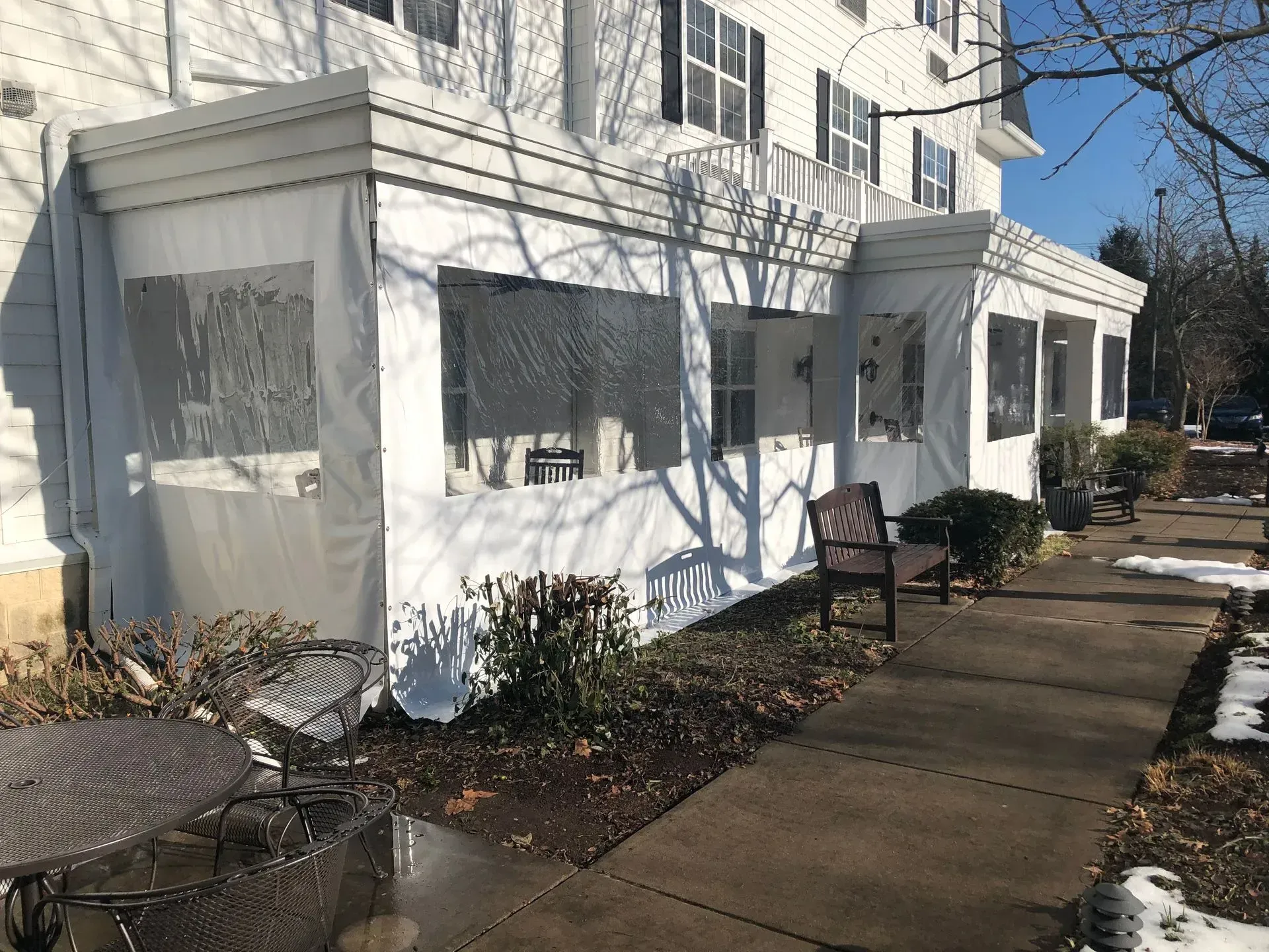 White apartment building entrance with boarded windows, sidewalks, benches, and bare shrubs in winter.