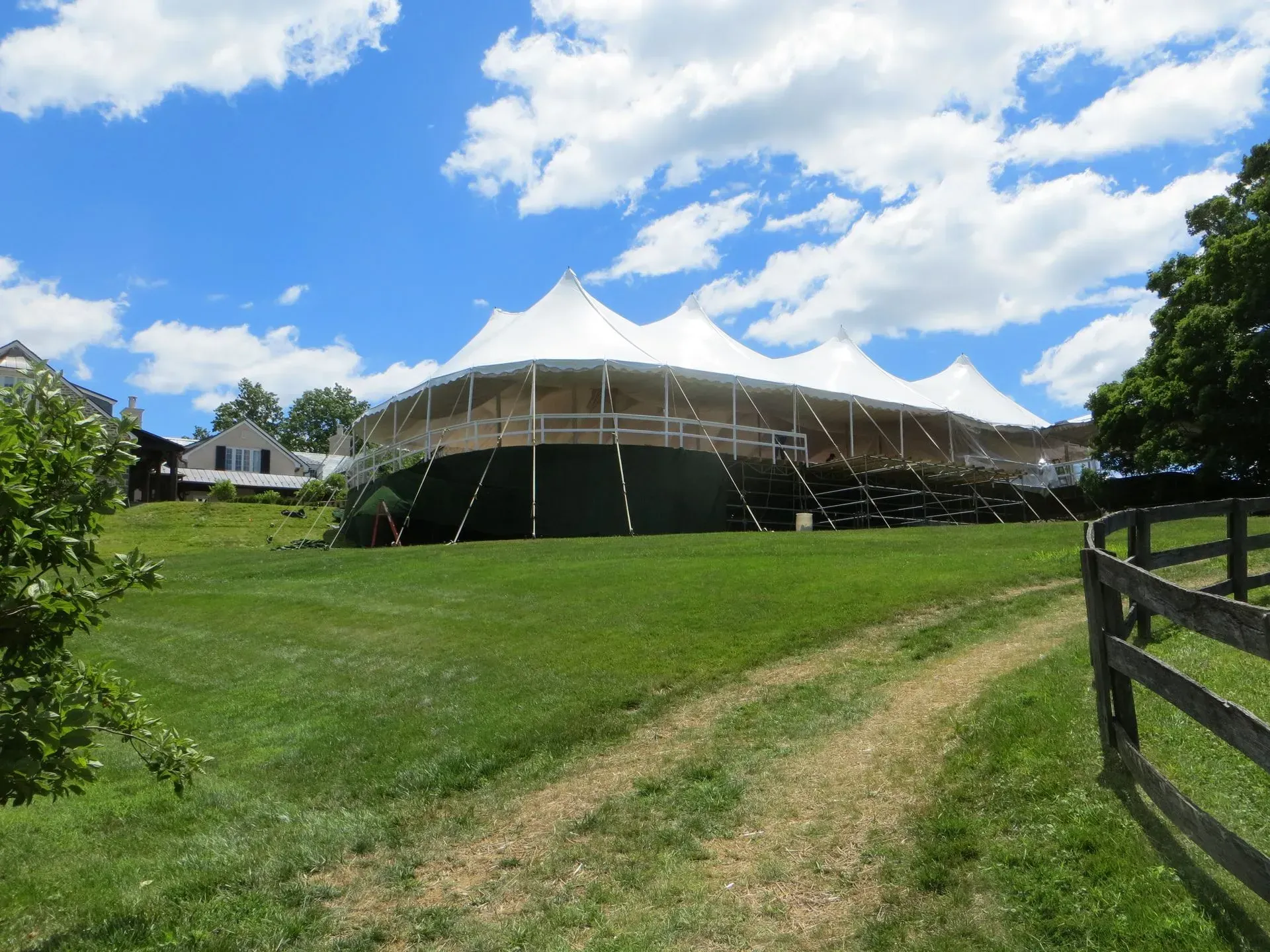 A large white event tent sits on a grassy field near a split-rail fence under a bright blue sky with scattered clouds.